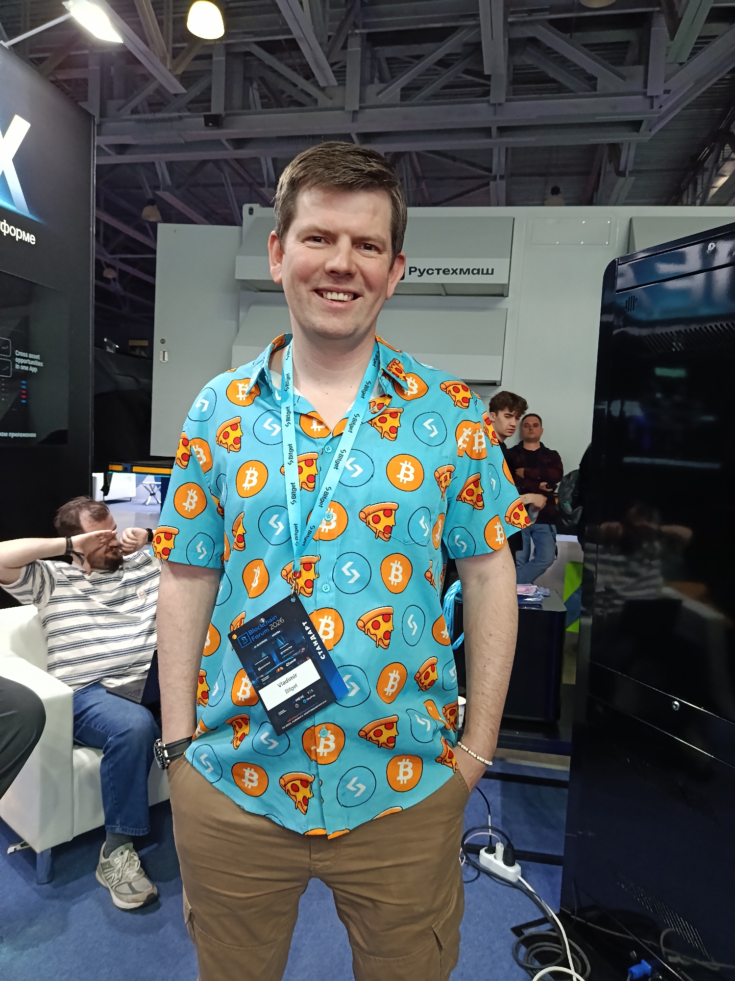 The image shows a smiling man in a light blue shirt with Bitcoin and pizza slice patterns. He is wearing a conference badge around his neck that reads "Blockchain Forum 2026" and "Vladimir Bitget." He is standing with his hands in his pockets.
Behind him is a crowd of people and equipment that reads "Pycтехмаш" (RusTekhmash). To the left, a man sits in a white chair covering his face. The image was most likely taken indoors at a convention or conference. This may be located in Moscow, Russia.
