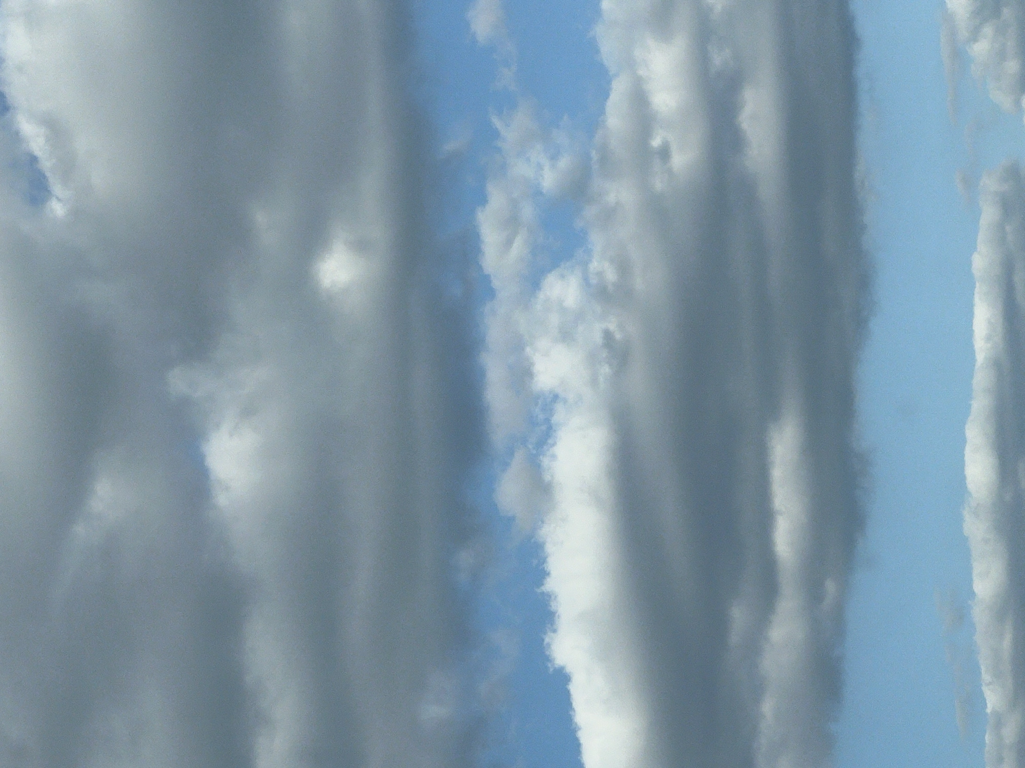 The image shows a cloudy sky above a body of water, likely a lake or river. The clouds are a mix of gray and white, with some areas of blue sky visible between them. The water appears calm, reflecting the colors of the sky. The scene suggests an outdoor setting in a natural environment, possibly a park or waterfront area. Without further context, it is impossible to determine the specific location within Warsaw, Poland.