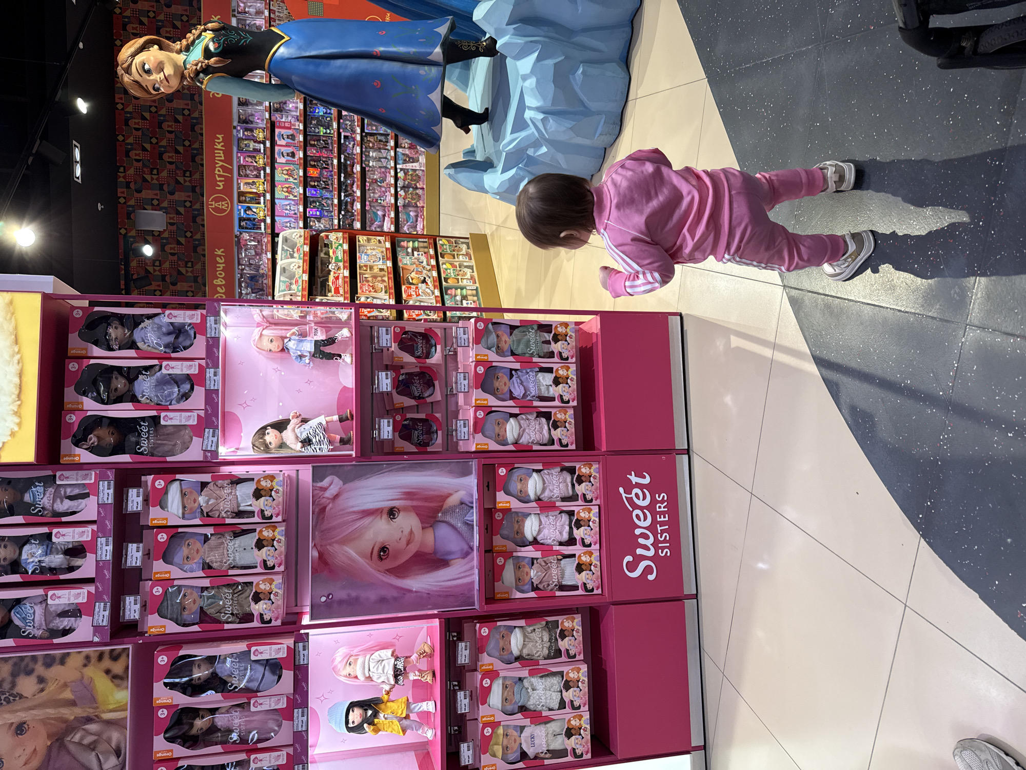 Here's a detailed description of the image:
**Scene Overview:** The photo appears to be taken inside a toy store, likely in a shopping mall, based on the floor tiles. A young child in a pink tracksuit is seen looking at a display of "Sweet Sisters" dolls. Above the doll display is a large display featuring Anna from Frozen.
**People:**
* **Child:** A young child is wearing a pink tracksuit and grey sneakers, lying on their stomach and looking at the doll display.
**Objects:**
* **Doll Display:** Features multiple rows of "Sweet Sisters" dolls in pink packaging. There are also large images of the dolls displayed on the cases.
* **Frozen Display:** A large plastic statue of Anna from the Disney movie "Frozen" is perched atop the doll display with a piece of blue ice.
* **Floor:** Tiled floor with a combination of dark grey and light beige-colored tiles.
* **Retail Shelves:** In the background, shelves are stocked with various other toys.
**Environment:**
* **Location:** The setting is a brightly lit, possibly indoor retail space. It's implied the location is Moscow, Russia based on OCR on the shelves in the background.
* **Lighting:** The scene is well-lit, casting distinct shadows.