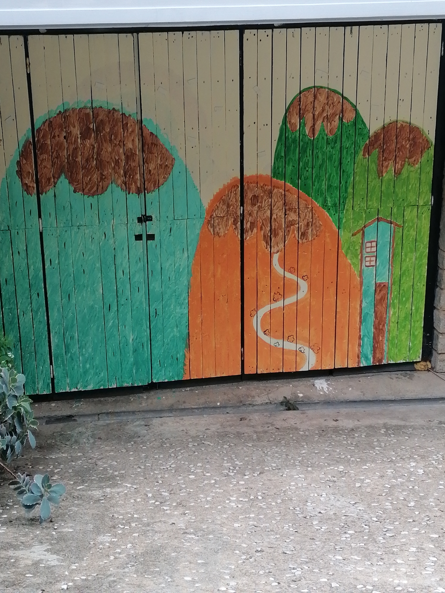 The image shows a section of a wooden gate painted with a whimsical landscape. The gate is divided vertically into two panels. The left panel is painted with a large, puffy brown shape that resembles a cloud, set against a turquoise-green mountain. The right panel features two green, cone-shaped mountains with brown tops, and a small blue house with a brown door and window frames. A winding white path with small, flower-like shapes is painted on an orange mountain on the far right. The gate is installed on a rough concrete ground. A small, green plant with succulent-like leaves is visible in the lower left corner of the image. The overall scene suggests a playful, artistic decoration. The specific location in Da Lat, Vietnam, cannot be determined from the image alone.