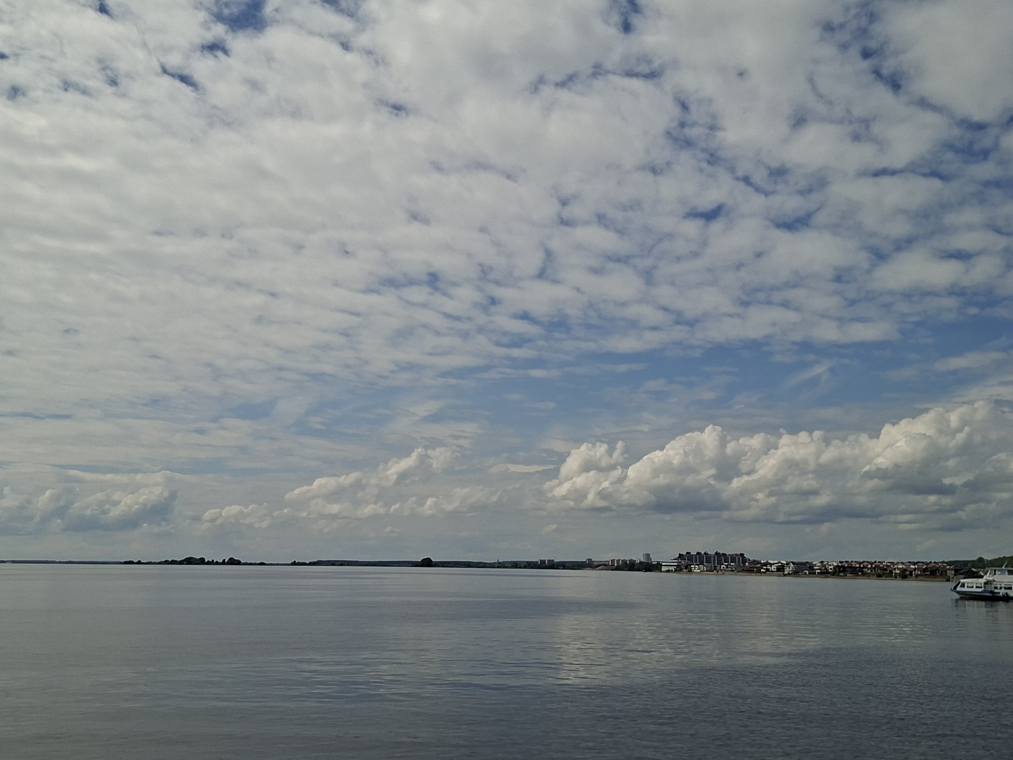 The image shows a wide, calm body of water, likely a river or bay, with a distant shoreline featuring a line of buildings and trees. The sky is filled with a mix of white, puffy clouds and lighter, wispy clouds against a blue sky. On the right side of the frame, a white boat with a dark hull is partially visible, moving across the water. The overall scene depicts a serene, overcast day with a city or town visible in the distance. This view is consistent with a location along the Volga River in Kazan, Russia, where residential areas and cityscapes can be seen across the water from various vantage points.