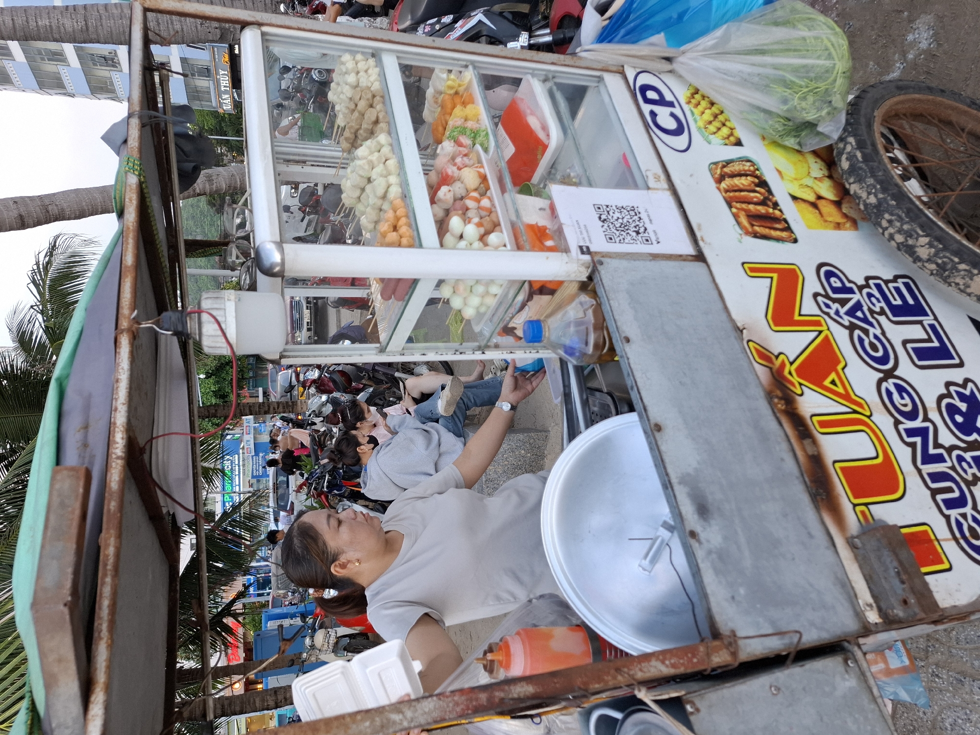 A woman is serving food from a mobile cart in what appears to be a street market in Bac Nha Trang Ward, Vietnam. The cart is loaded with various food items, including skewers of meatballs, sausages, and other fried goods displayed in a glass-fronted cabinet. The woman is wearing a gray t-shirt and is holding a white take-out container. Behind her, several people are seated on a stone bench, and numerous motorcycles are parked. The background shows palm trees, buildings, and a hazy sky, suggesting an outdoor, urban setting. The cart itself is adorned with colorful lettering and images of food.
