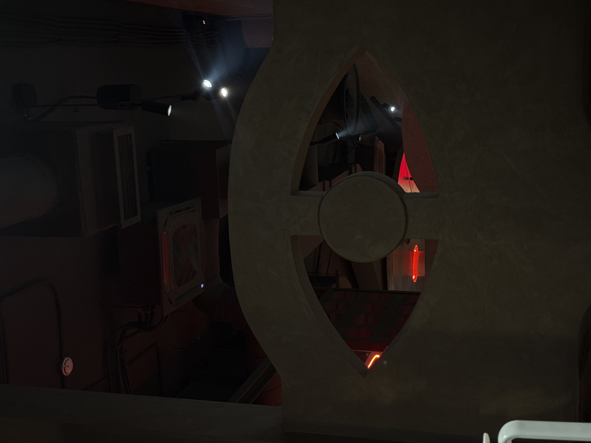 The image shows an interior scene with a dark, moody ambiance, likely a restaurant or bar. In the foreground, a large, curved concrete structure with an eye-shaped cutout dominates the frame. Through the cutout, glimpses of red lighting and what appear to be ceiling fixtures are visible. The ceiling above is partially visible, showing electrical conduits, a smoke detector, and HVAC units. A few track lights are also present, casting bright spotlights. The overall impression is one of modern, industrial-inspired decor with dramatic lighting. The location in Borodino, Russia, is not visually discernible from the image itself, as there are no specific architectural features or signage that would indicate this location.