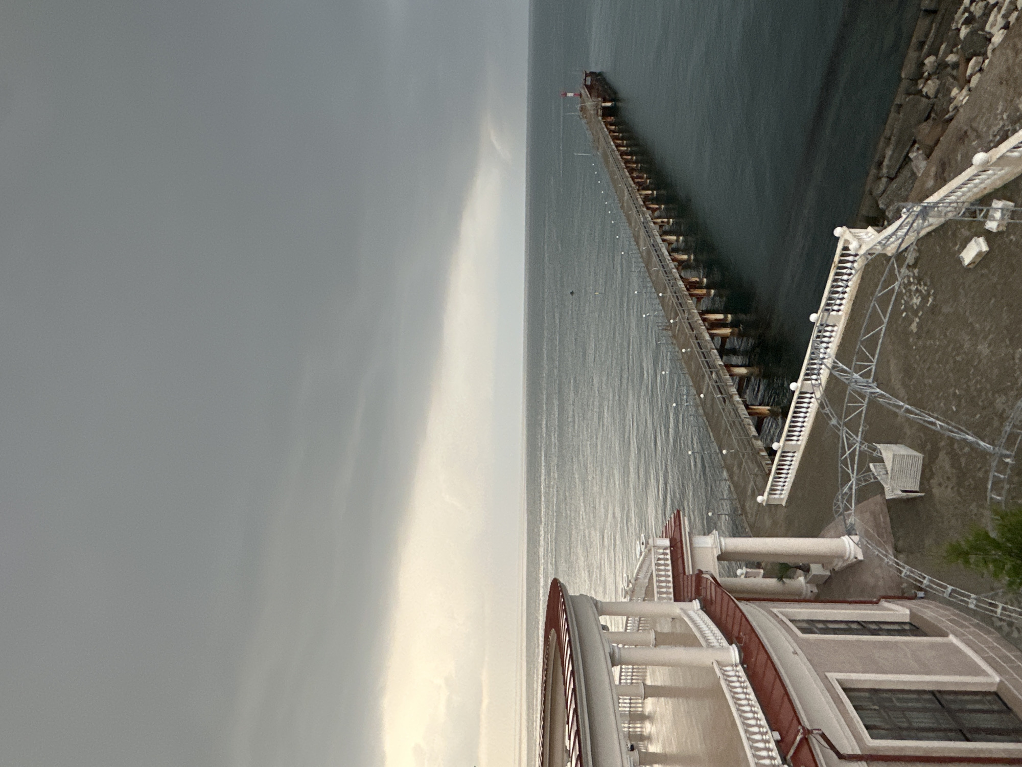 An outdoor, elevated, eye-level, wide shot captures a cloudy, overcast day at a coastal location. The scene is dominated by a vast expanse of dark, calm water, stretching to a horizon where the sky meets the sea. A long, wooden pier juts out into the water from the right side of the frame, lined with lampposts and terminating in a lighthouse at its end. In the foreground, to the left, a portion of an ornate, curved building is visible, featuring white columns, a red-tiled roof, and a white balustrade overlooking the water. A paved area with some metal structures is situated below the building's balcony. The overall atmosphere is somber and quiet, with no people or animals present.