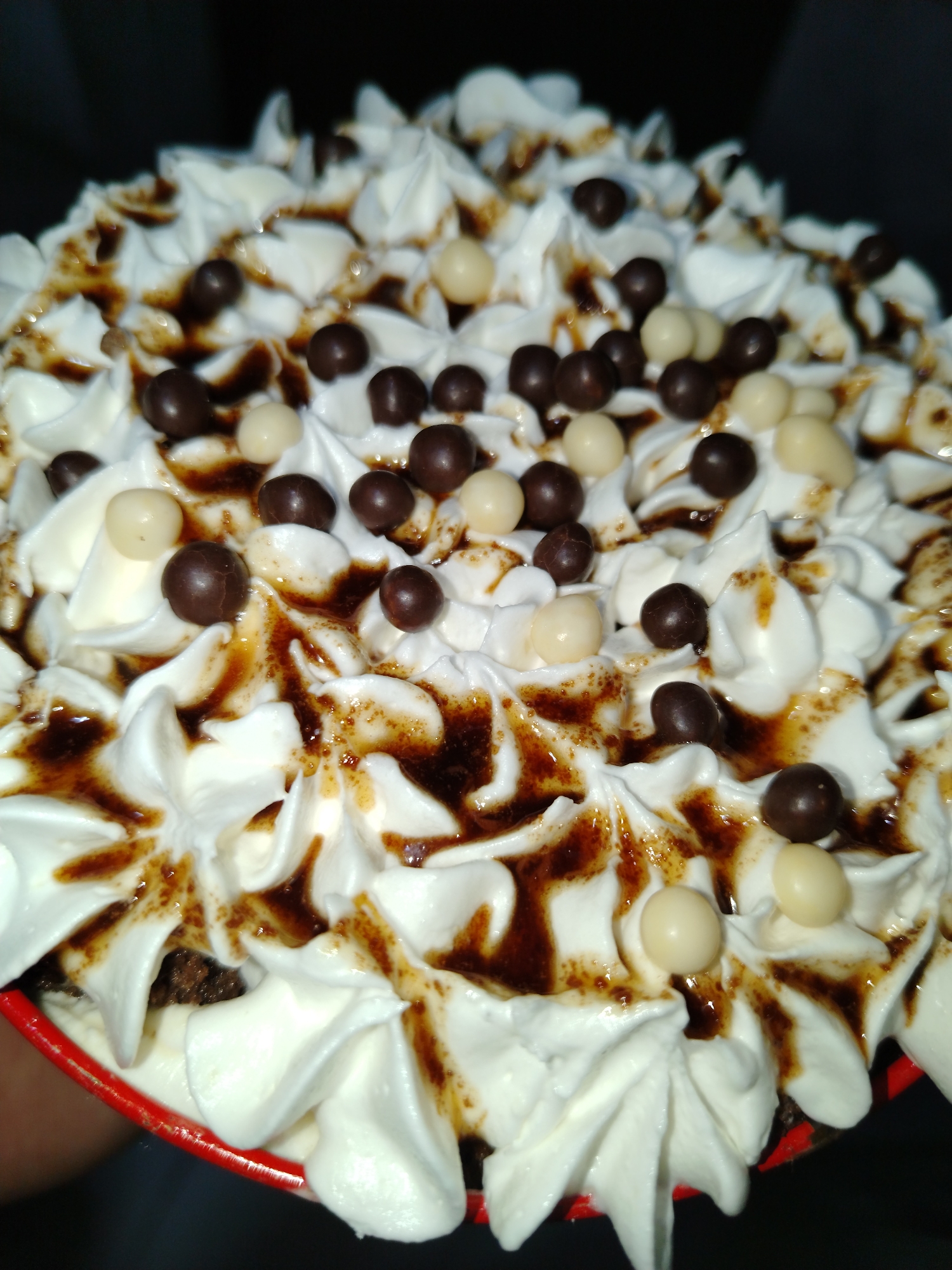 The image shows a close-up of a dessert, possibly a cake or cupcake, topped with piped white frosting. The frosting is drizzled with a dark brown sauce, and decorated with small, round chocolate and white candy balls. The dessert is in a red cup or container. The location is Tinaquillo, Venezuela.