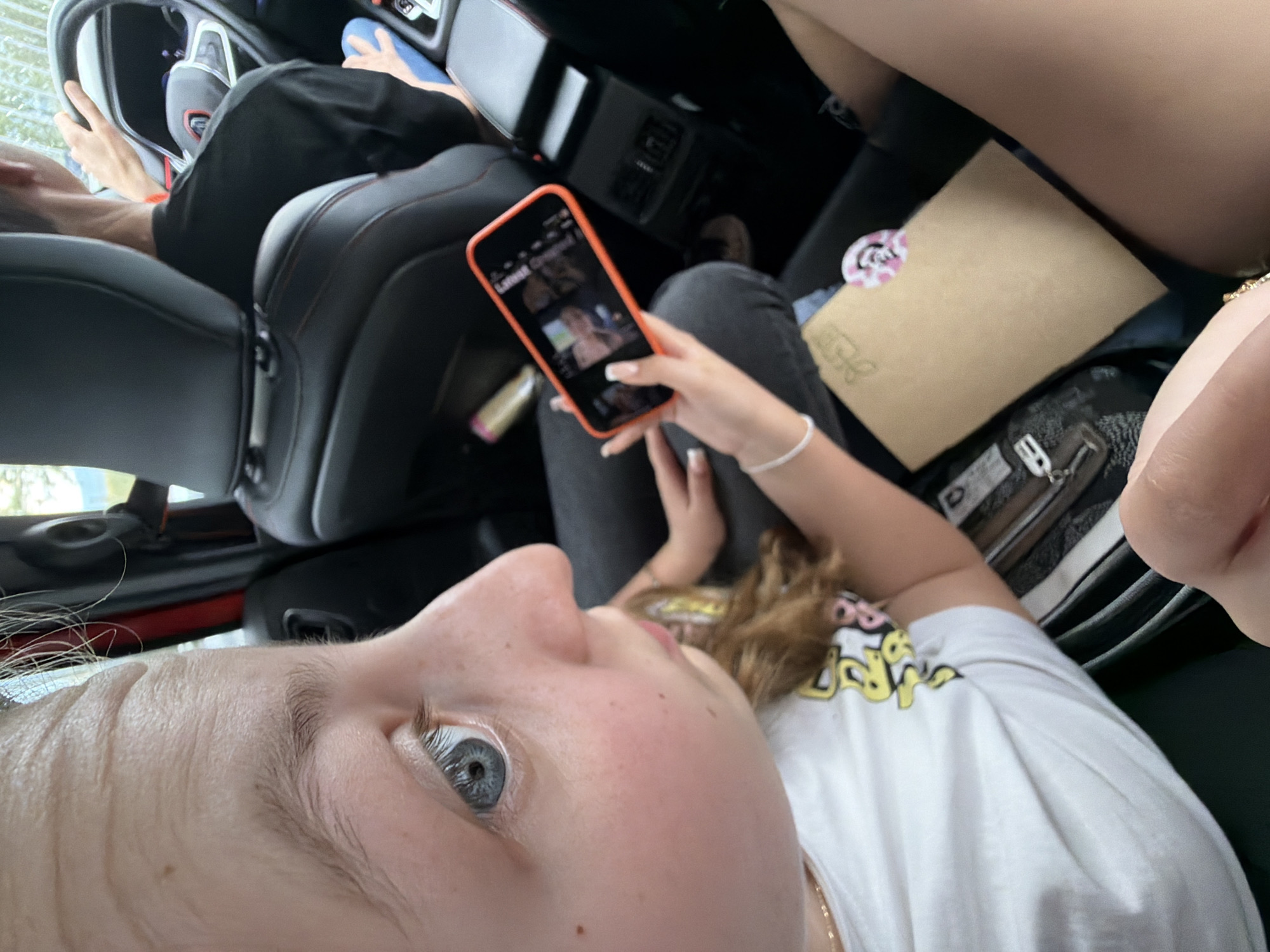 A person is taking a selfie while sitting in a car. The person is holding a cell phone in their right hand, looking at the screen. Another person is driving the car. There is a purse and an envelope on the passenger seat. The location is indeterminate, but the city is Chokoba, Bulgaria.