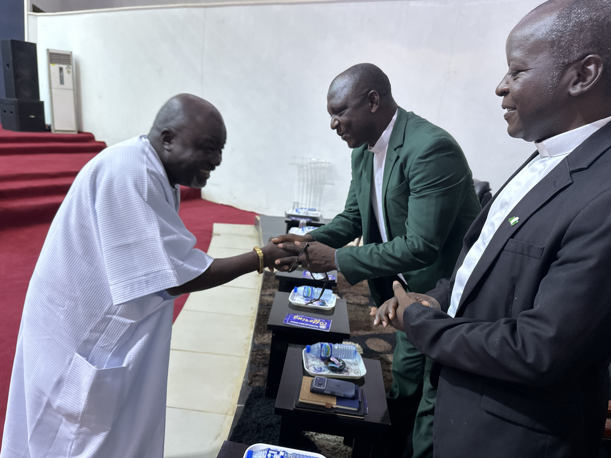 In a dimly lit indoor space, possibly a meeting hall or conference room in Television, Nigeria, three Black men are engaged in a handshake. The man on the left, wearing a white robe with a textured pattern and a gold bracelet on his left wrist, is shaking hands with the man in the middle, who is dressed in a dark green suit over a white collared shirt. The man on the right, viewed from the side, wears a black suit jacket over a white clerical collar and shirt, with a small Nigerian flag pin on his lapel. He is also reaching out his hand. The handshake between the first two men appears firm and is the focal point of the interaction.
In the foreground, a series of low, dark rectangular tables are arranged. On these tables are various items, including small bottles of water on decorative plates, eyeglasses, cell phones, and what appear to be booklets or notebooks. A purple card with white lettering, possibly an event program or name tag, is visible on one table. The floor is covered with a patterned carpet. In the background, a white wall provides a backdrop, and to the left, a red carpeted incline leads upwards, with two black audio speakers and a white air conditioning unit on a platform. The lighting is subdued, casting shadows and highlighting the central figures and the handshake.