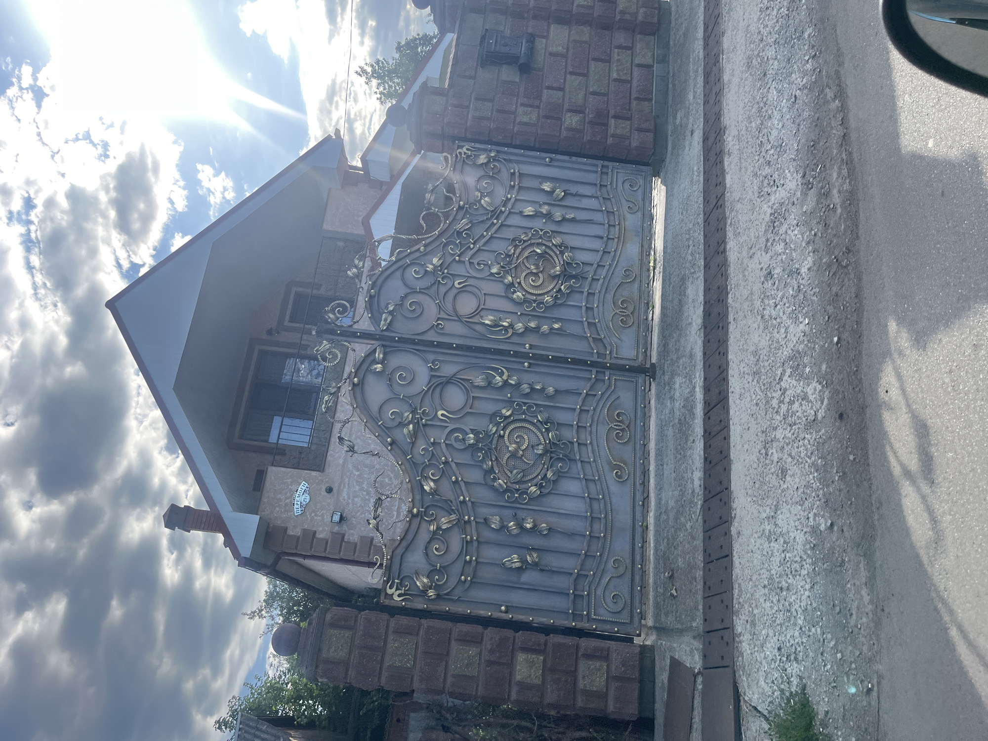 This image captures a street view in Storozhnytsia, Ukraine, featuring a large, ornate metal gate in front of a house. The gate is predominantly gray with intricate gold scrolling and leaf-like embellishments, divided into two sections. It is flanked by brick pillars, with the left pillar appearing to be part of a larger fence. The house behind the gate has a light-colored facade with brick accents and a red roof. A chimney is visible on the roof. The sky above is partly cloudy with bright sunlight filtering through, creating a lens flare in the upper right portion of the frame. The ground in front of the gate is paved with concrete and a section of asphalt road is visible in the foreground, with a car's side mirror also visible on the lower right. A small sign with Ukrainian text is affixed to the house above the gate.