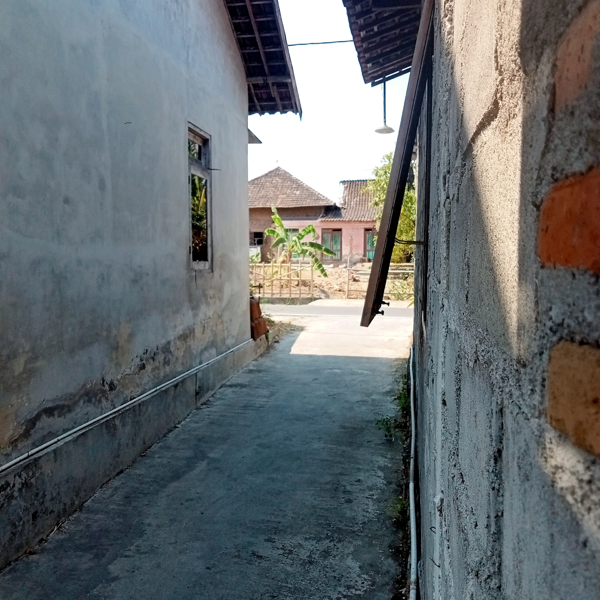 The image depicts a narrow alleyway between two buildings, suggesting a residential area in Jaten, Indonesia. The building on the left has a rough, light gray plastered wall with a visible crack and a small, open window. A white pipe runs along the base of this wall. The building on the right is constructed of brick and has a rough gray plastered exterior, with visible orange bricks in some areas. A dark, wooden plank is attached to the right wall at an angle, extending into the alley. Above, the roofs of the buildings are visible, with exposed wooden beams and tiling. In the background, through the alley, several houses with tiled roofs are visible. One house has a pink facade and green wooden shutters. A banana tree stands in front of a bamboo fence. The ground of the alley is paved with concrete, showing some wear and tear. The sky is bright and clear, indicating daytime. The overall impression is one of a simple, possibly rural or semi-urban, Indonesian settlement.