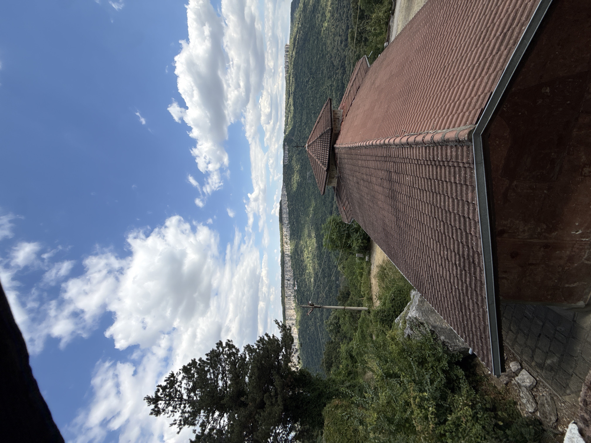 The image shows a view of a mountainous landscape with a building in the foreground. The building has a red tiled roof and appears to be part of a religious complex, possibly a monastery, given the small cross visible on the roof. The landscape is characterized by lush green vegetation covering the slopes of the mountains. A prominent cliff face runs across the horizon, dotted with structures that suggest ancient settlements or architectural remnants. The sky is blue with scattered white clouds. The scene suggests a remote and possibly historical location. This location is the Rock-hewn Churches of Ivanovo, near the village of Ivanovo, in the Ruse Province of Bulgaria. Samovodene is a village in the Veliko Tarnovo Province, not Ruse.