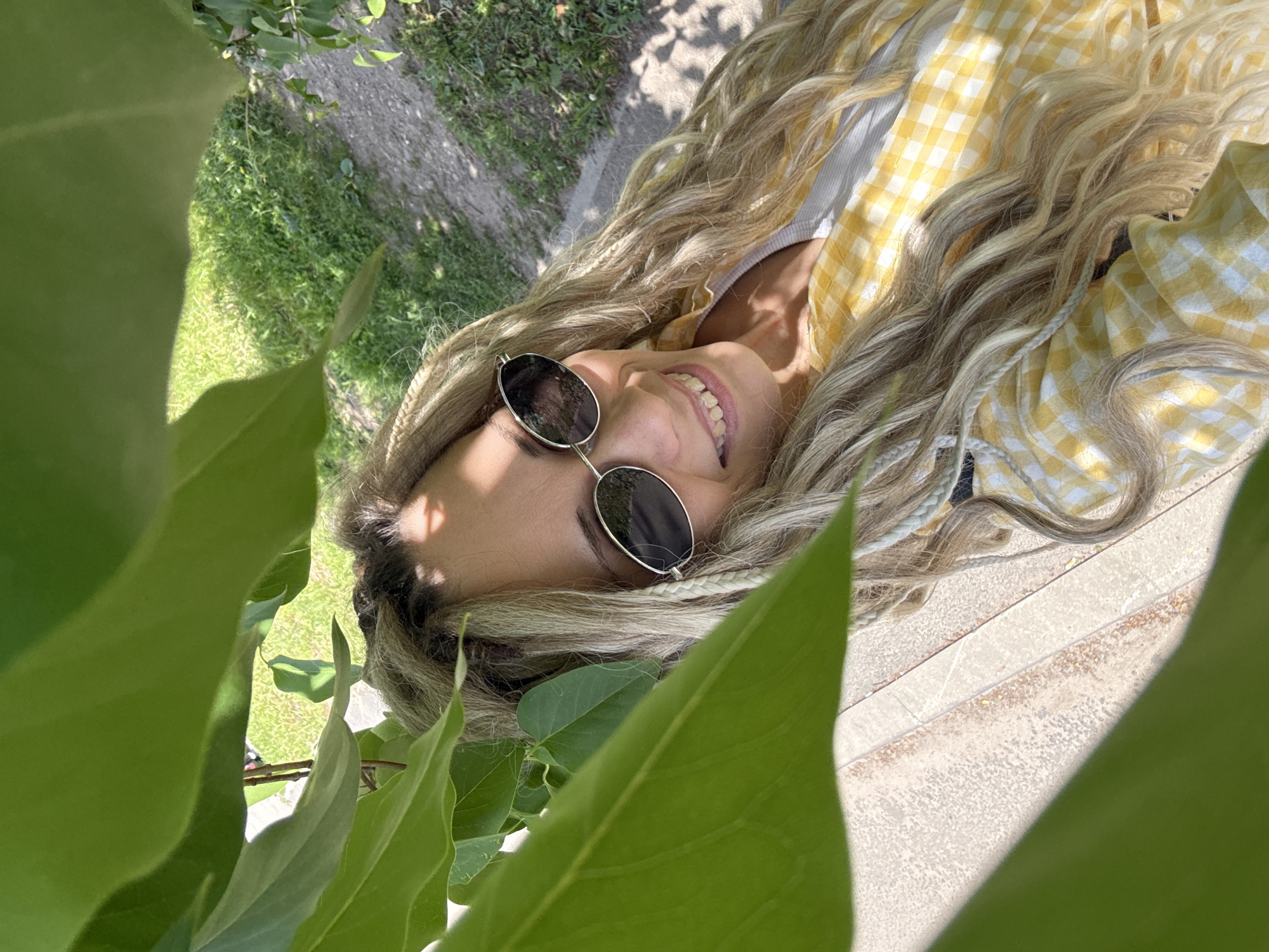 A young woman is captured in an outdoor, upward-facing portrait, framed by large, vibrant green leaves in the foreground. Her head and upper torso are visible, with her long, wavy, blonde hair cascading around her. Several thin braids are interwoven into her hair. She is wearing round-framed sunglasses with dark lenses and a cheerful yellow and white gingham-patterned top, under which a white or light-colored striped garment is visible.
The woman is smiling broadly, revealing her teeth, and appears relaxed and content. The bright sunlight casts strong shadows, indicating a sunny day, likely during midday or early afternoon. She seems to be lying on her back or reclining on a light-colored surface, possibly concrete or pavement, which is visible to the right and below her head. In the background, beyond the foliage and the surface she rests on, there is green grass and a winding dirt or gravel path, suggesting a natural setting such as a park or garden. The overall impression is one of leisure and enjoyment of the outdoors. There is no visible text in the image.