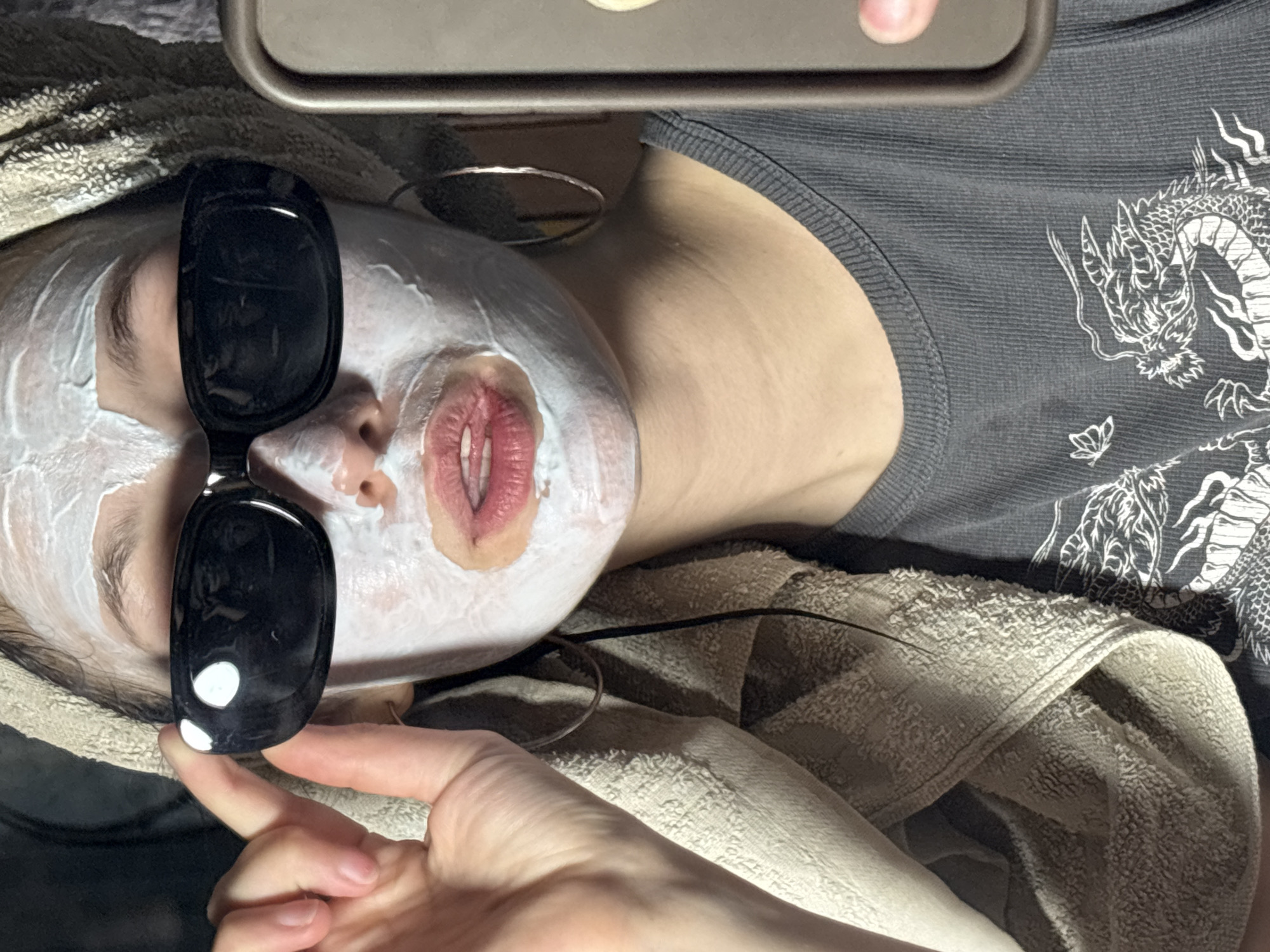 A person with a white facial mask and sunglasses is lying down. The person is wearing a gray shirt with a white dragon design on it. A tan-colored towel is wrapped around the person's head. A hand is holding the sunglasses. A metal tray is visible at the top of the frame. The location is unknown and cannot be determined from the image.