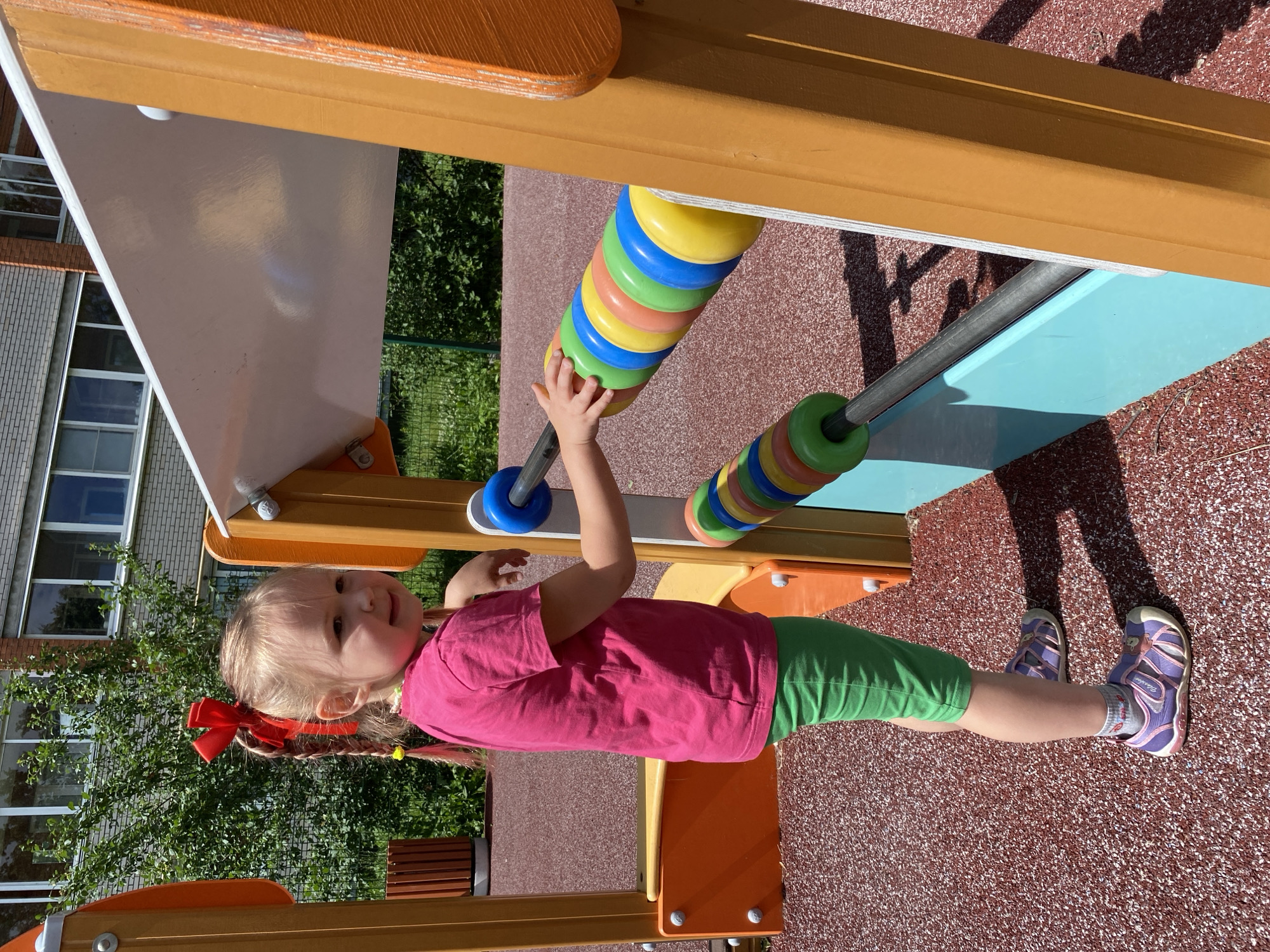 A young blonde girl with pigtails adorned with a red bow stands on a playground. She wears a pink t-shirt and green shorts, with purple and pink sandals on her feet. The girl is positioned on a red, textured surface, which appears to be part of the playground. In front of her, a colorful abacus with large, segmented beads in yellow, blue, green, and orange is mounted horizontally. Her right hand is reaching towards the abacus.
Behind the girl, a building with brickwork and windows is visible, partially obscured by green foliage. The playground structure itself is made of brightly colored materials, including orange, yellow, and turquoise elements. Sunlight casts shadows on the playground, suggesting it is daytime, likely sunny. The overall scene depicts a playful moment for a child on an outdoor playground in Chaikovsky, Russia.