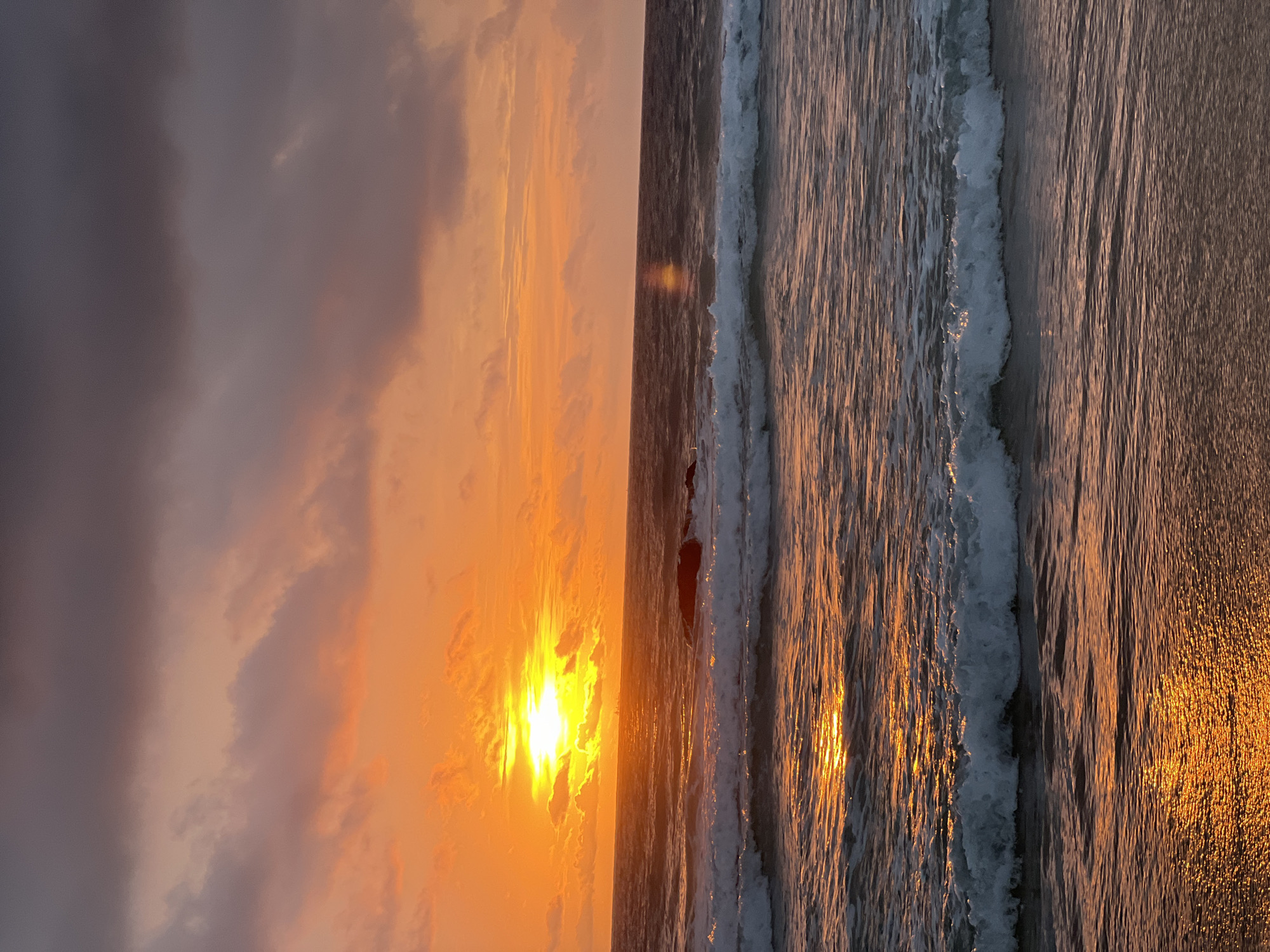 A vibrant sunset paints the sky in hues of orange and pink over the Andaman Sea. The sun, a bright orb, dips towards the horizon, casting a golden reflection on the undulating waves. Dark, moody clouds drift above, contrasting with the fiery sky. In the middle ground, a dark, rocky outcropping emerges from the water, its surface wet and glistening. White-capped waves roll towards the shore, their foamy crests illuminated by the setting sun. The foreground shows the gentle lapping of water against a sandy beach, with the warm tones of the sunset creating shimmering patterns on the wet sand. This scene is likely captured at a beach in Choeng Thale, Thailand, known for its stunning sunsets. There are no people or animals visible in this image.