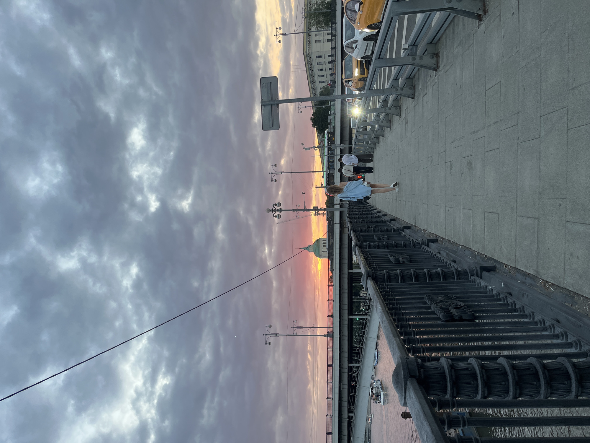 The image depicts a sunset scene on a bridge in Moscow, Russia. A woman in a blue shirt and shorts walks away from the camera on the pedestrian walkway. Several people are walking ahead of her, and cars are driving on the road to her right. A streetlamp stands on the sidewalk, and a wire stretches across the sky. In the background, a domed building is visible against a sky filled with dramatic clouds tinged with pink and orange from the setting sun. Boats are on the river below, and a bridge spans across it. The scene appears to be on the Patriarchal Bridge or Krymsky Bridge due to the architectural style of the railing and the visible domed building.