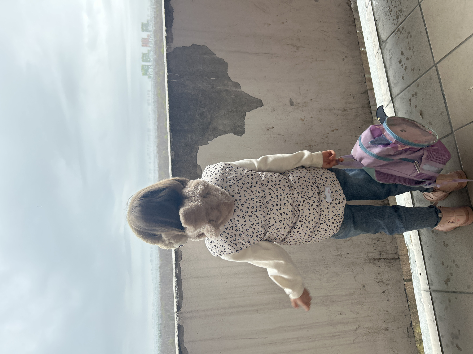 A young girl with light brown hair wearing a patterned jacket, jeans, and pink shoes stands next to a concrete wall. She holds a purple backpack. The backdrop shows an overcast sky and glimpses of distant buildings. The location is Moscow, Russia.