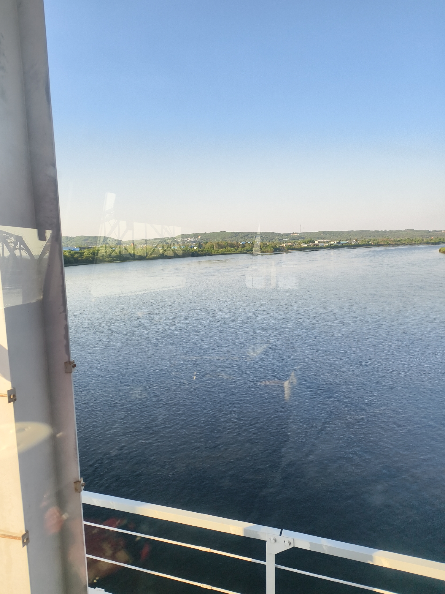 The image shows a wide river view from an elevated platform, possibly a bridge or a high building, as suggested by the white railing and structural support visible in the foreground. The river is wide and calm with subtle ripples on its surface. On the distant bank, there is a stretch of land covered with greenery, including trees and some low-lying buildings. The sky is clear and blue.
There is no reference to Bardagon, Russia in the image itself. It may be a part of the image description. No people or actions are visible. The primary objects are the river, the land, the sky, the railing, and the building support. The interaction is between the natural environment and the man-made structures. The environment is overall bright and open.