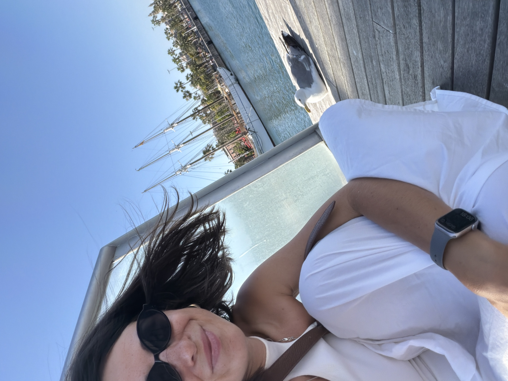 A woman wearing sunglasses, a white top, and white pants is posing for a selfie on a wooden pier in Barcelona, Spain. Her dark hair is windswept, and she is smiling. To her right, a seagull is resting on the wooden planks. In the background, a large white sailboat with tall masts is docked in the blue water, with palm trees and buildings lining the waterfront. The sky is clear and blue. The woman is wearing a watch on her left wrist.