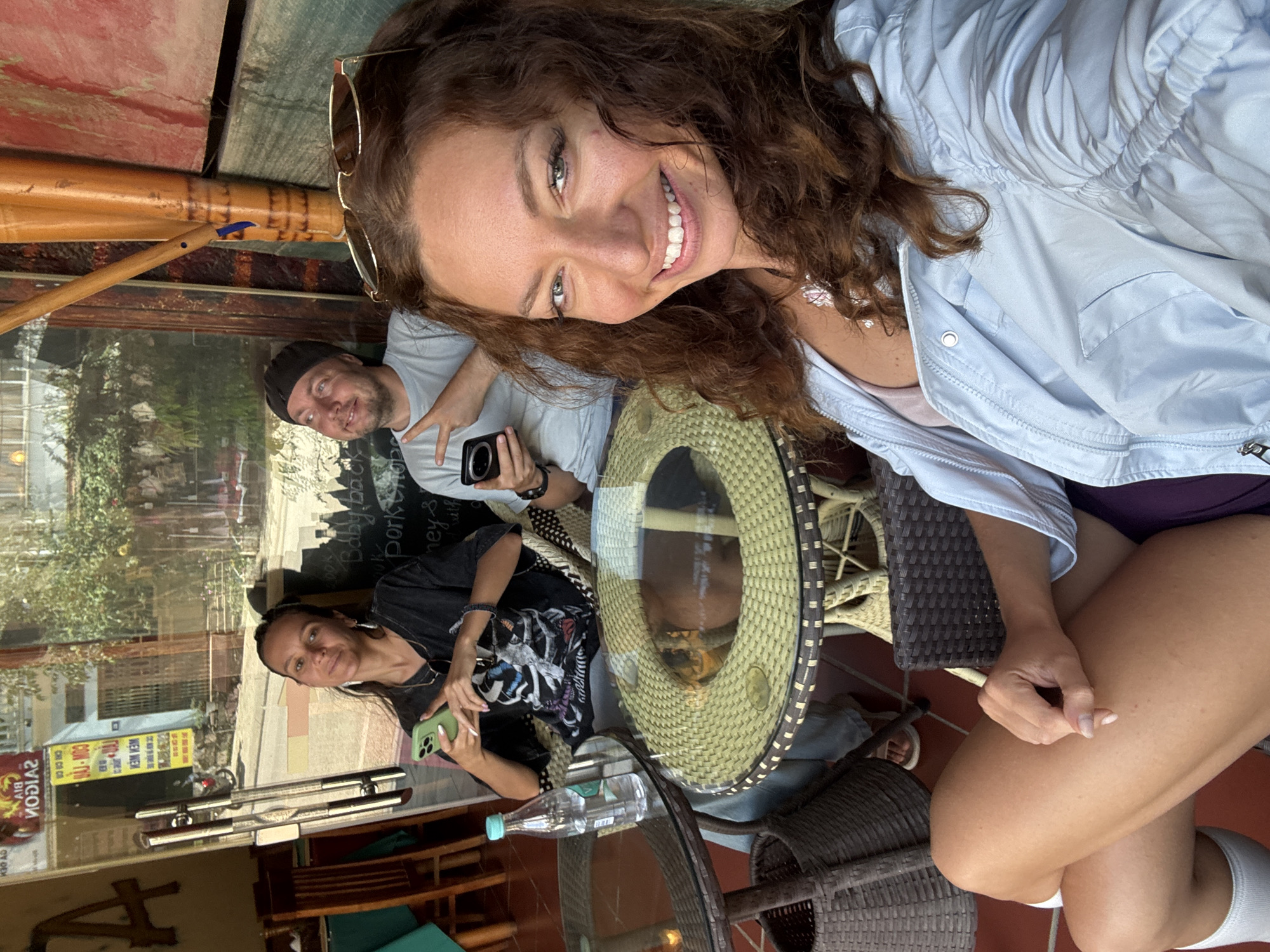 The image depicts three people seated at tables outdoors in what is identifiable as the city of Đồng Văn Commune, Vietnam, as evidenced by signage in Vietnamese visible through the glass of a nearby storefront.
The predominant individual in the foreground is a woman with light skin, brown curly hair, and sunglasses perched on her head. She is wearing a light blue jacket and is smiling broadly at the camera. Seated across from her is another woman with dark hair, wearing a black t-shirt and holding a cell phone with a green case. Behind them is a man wearing a black hat, a grey t-shirt, and holding a camera.
The group is seated around two tables; one table is made of glass with a wicker base, and has a clear plastic water bottle on it, and the other is made of wicker. The tables are arranged in an outdoor area that appears to be part of a café or restaurant.
Through the glass of the storefront, there is a glimpse of the street scene with signs in Vietnamese, which identify the location as Đồng Văn Commune, Vietnam. The overall setting suggests a casual social gathering or meal taking place outdoors at a local establishment.
