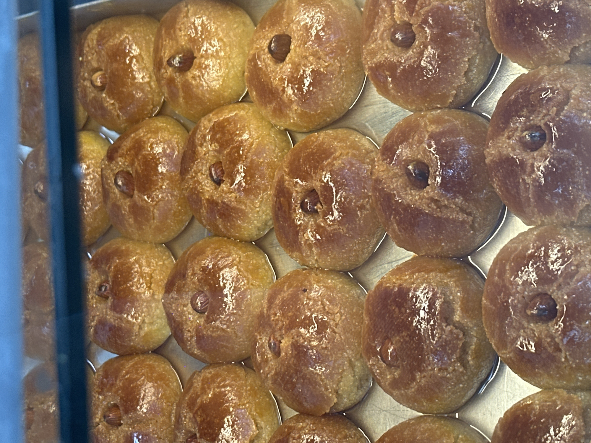 The image displays a close-up, eye-level view of a tray filled with numerous, golden-brown, round pastries. Each pastry is topped with a single nut, possibly an almond or hazelnut, embedded in the center. The pastries appear to have a slightly flaky texture and are arranged in neat rows within a metallic tray. The lighting creates highlights on the glazed surface of the pastries. The background is blurred, but suggests a retail environment, possibly a bakery or pastry shop. There is no information in the image to suggest a location in Amsterdam, Netherlands.