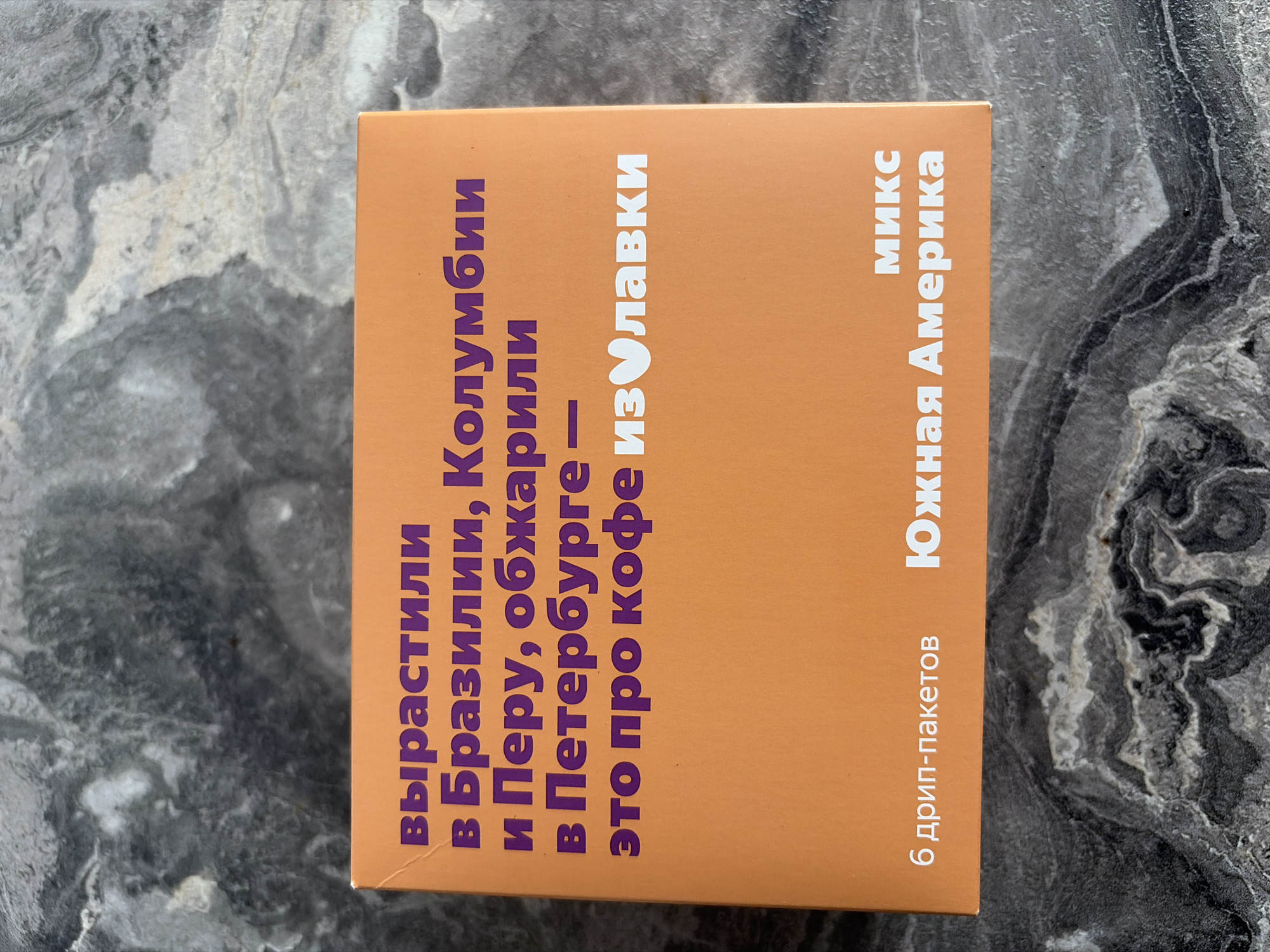 The image shows a close-up, top-down view of an orange box with text printed in purple and white on a dark, textured surface. The text on the box is in Russian. The purple text on the top part of the box reads "вырастили в Бразилии, Колумбии и Перу, обжарили в Петербурге – это про кофе из Улавки". The white text at the bottom of the box reads "6 дрип-пакетов" on the left and "МИКС Южная Америка" on the right. There are no people or identifiable landmarks present in the image. The image does not provide any information about the city of Sochi, Russia.