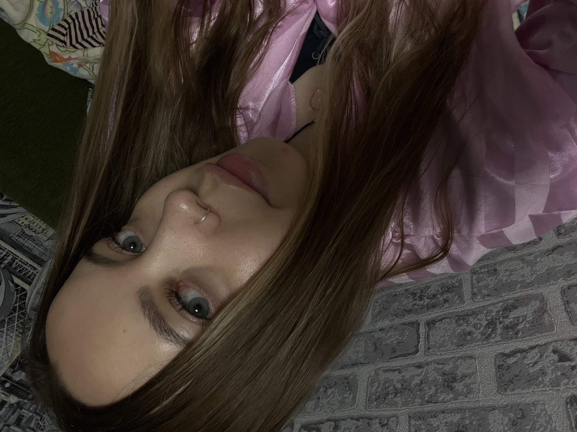 A close-up, angled view of a person lying down. The person has light skin, light eyes, brown hair, and a nose ring. They are wearing a pink garment. The background appears to be a textured wall, possibly wallpaper, resembling gray bricks. Other surfaces are seen in the background as well, one of which looks like a pillow. The location is not discernible from the image.