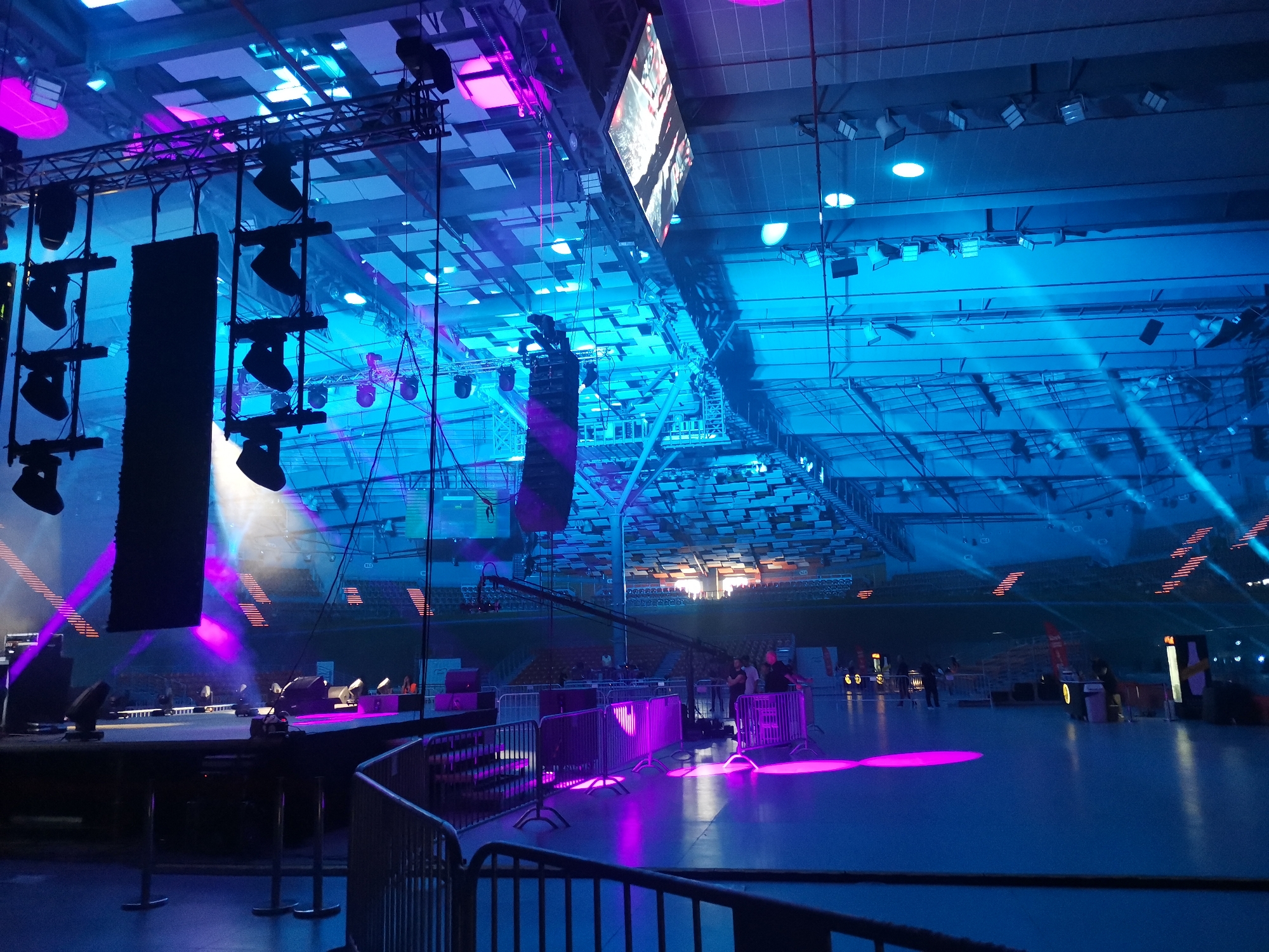 This detailed image depicts the interior of the Kolodruma Sports Hall in Plovdiv, Bulgaria, configured for a large-scale event. The expansive multi-purpose arena is bathed in striking stage lighting, primarily cool blue and vibrant magenta.
A prominent stage structure is positioned on the left, equipped with multiple suspended black line array speakers and an intricate arrangement of spotlights affixed to metal trusses. These lighting fixtures project intense beams of blue and magenta light across the floor and towards the ceiling. Overhead, a large, diamond-oriented LED screen is suspended from the high, industrial-style ceiling, displaying bright, dynamic digital content. The ceiling itself features a visible grid of trusses, ventilation ducts, and a variety of square and circular recessed lights, some emitting white light, others contributing to the blue ambient illumination.
The vast, smooth floor surface, appearing light grey or blue, is largely empty and reflects the magenta and blue stage lights in distinct pools. In the mid-ground, grey metal crowd control barriers are strategically placed, defining areas around the stage and potential audience zones. A long camera jib arm extends from the right mid-ground towards the stage, indicating professional video recording capabilities.
A small group of approximately three to five individuals, likely event personnel, are observed standing in the mid-ground to the right, near the barriers, appearing to be engaged in conversation or overseeing preparations. In the background, tiered spectator seating areas are visible, with some sections illuminated in a warm orange-brown hue. Additional indistinct figures and vertical signage or display structures are discernible further back, contributing to the event setup. The overall scene suggests a significant public event is in the final stages of preparation.