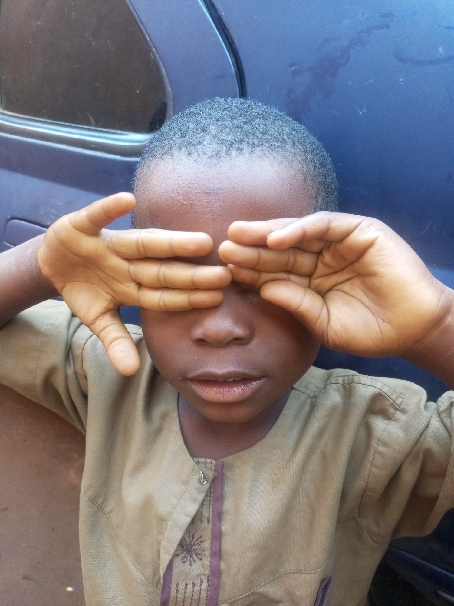 A young boy, appearing to be of Nigerian descent, is depicted in a close-up, head-and-shoulders shot. He is wearing a light brown, traditional Nigerian garment with dark purple embroidery on the front. His hands are placed over his eyes, partially obscuring his face, with his fingers spread. The boy's skin tone is dark brown, and his hair is short and dark.
The background is predominantly a dark blue surface, likely the side of a vehicle, with a dark, reflective car window visible to the left of the boy's head. The overall lighting suggests an outdoor setting, possibly during daylight. The boy is positioned in what is described as the city of Tempure, Nigeria.
