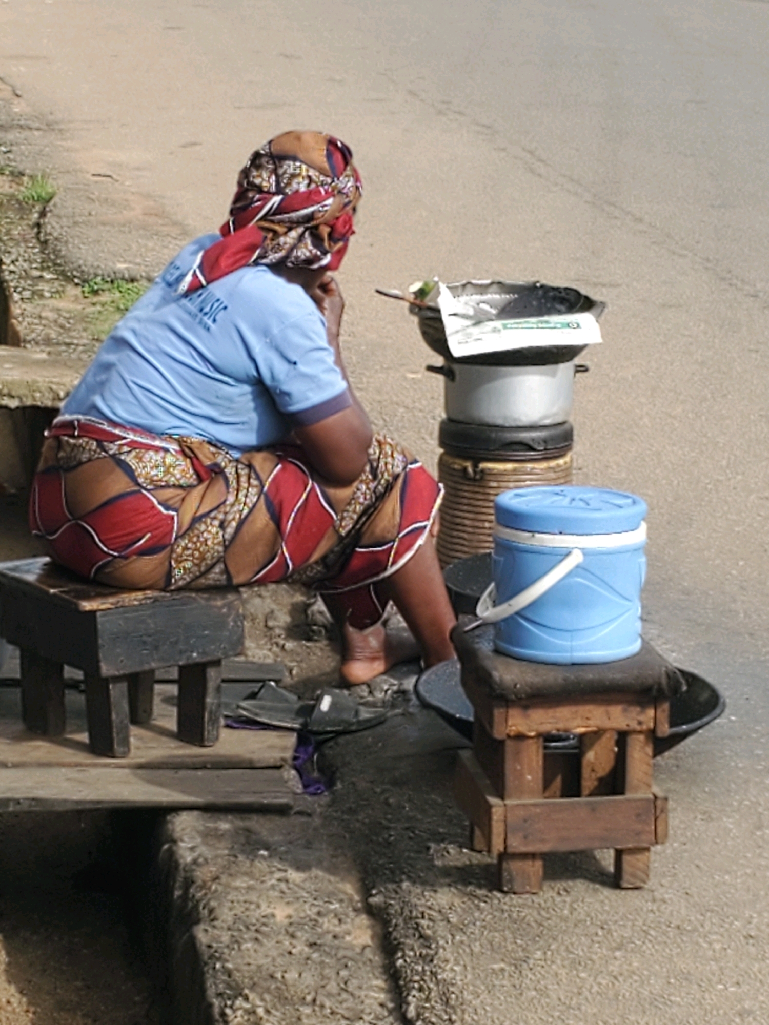 A woman, wearing a blue t-shirt and a colorful patterned wrap, sits on a wooden stool beside a road in Jos, Nigeria. She has her headscarf covering her head and a portion of her face. Next to her, on a slightly taller, stacked set of containers, is a large black wok, likely for cooking. In front of the woman, on another wooden stool, is a blue and white cooler. The ground is paved with asphalt.