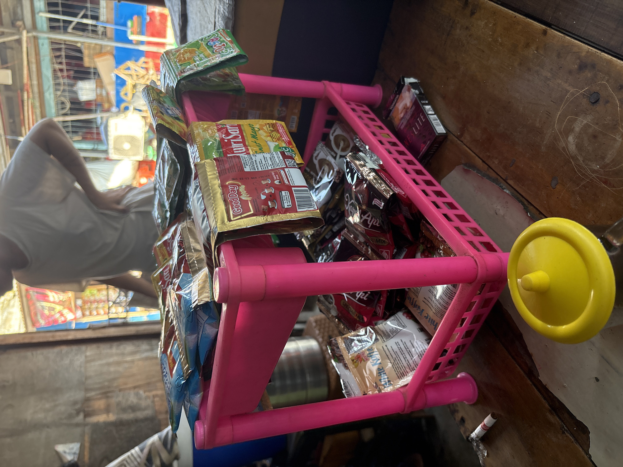 The image depicts a close-up view of a small, likely outdoor, convenience store or roadside stall, possibly located in North Jakarta, Indonesia. The central focus is a bright pink, two-tiered plastic shelf holding various packaged food and beverage items. The top shelf is laden with small, colorful packets of what appear to be coffee or powdered drink mixes, including brands like "Good Day" and "NutriSari" with fruit flavors like "Sweet Mango". Some packets are stacked and others are hung from the shelf.
The lower shelf of the pink unit contains more packaged goods, including what seem to be chocolate bars or similar confectionery. To the right of the lower shelf, a pack of cigarettes labeled "NES LITE" is visible. In the foreground, to the left of the shelf, a partially smoked cigarette lies on a wooden surface. A yellow, round plastic lid sits on the wooden table in the lower-right portion of the image.
In the background, a person wearing a white t-shirt is partially visible, with their back to the camera. They appear to be standing behind the stall. The stall itself seems to be constructed with rough materials, possibly corrugated metal or wood, suggesting a humble setting. Beyond the stall, colorful decorations or products are visible, hinting at the lively atmosphere often found in such local markets. The overall lighting suggests it is daytime, with sunlight illuminating the scene.