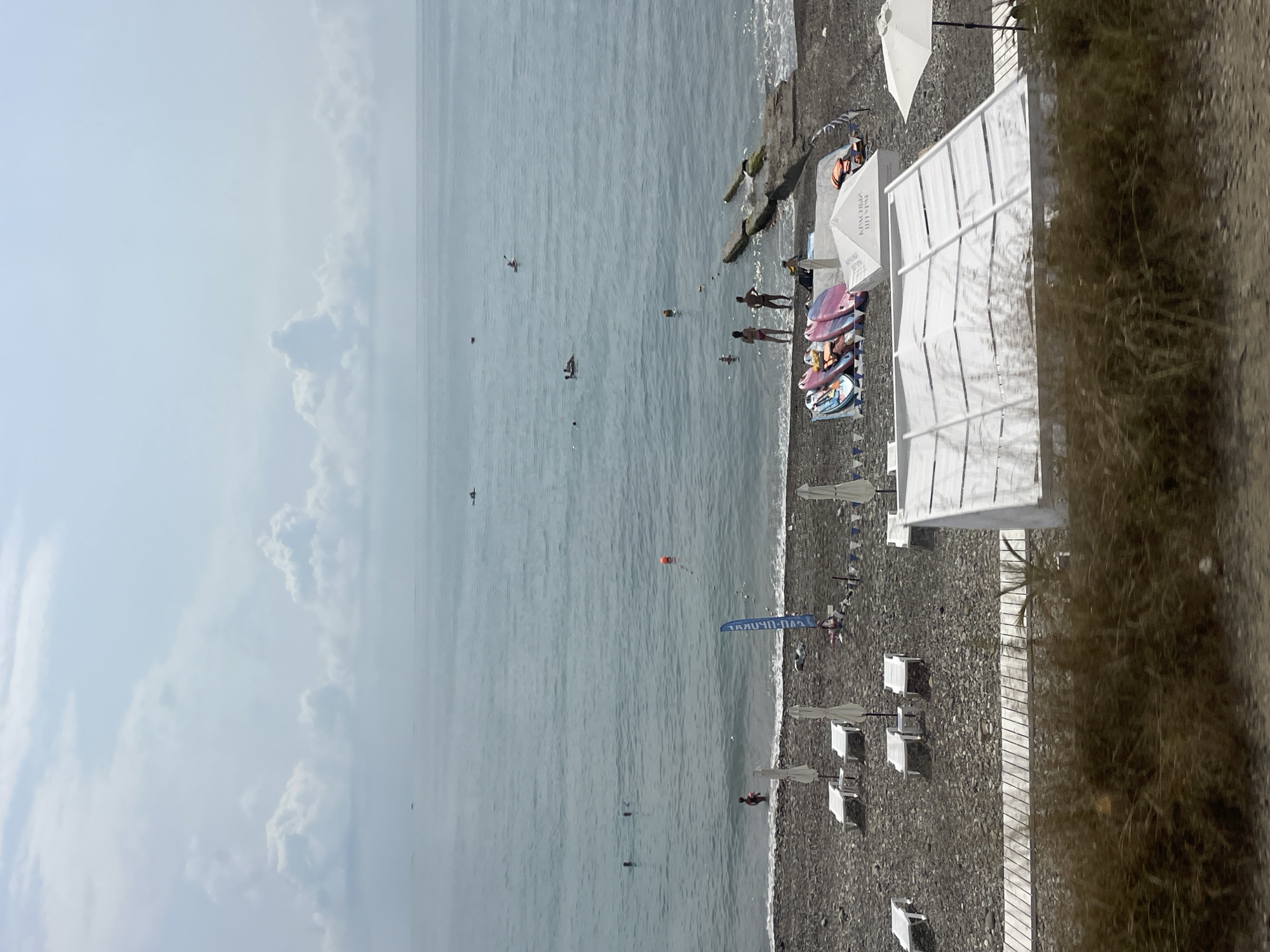 This is an eye-level, medium shot of a pebbled beach on a cloudy day. The ocean occupies the upper two-thirds of the frame, with several people swimming and two kayaks visible on the calm water. A red buoy floats in the water near the shore. On the beach, there are several white lounge chairs and umbrellas, some arranged on a wooden walkway. A row of surfboards is propped up on the sand, and a white tent structure stands prominently in the foreground, with another smaller tent structure visible behind it. A concrete pier extends into the water on the right side of the image. The scene appears to be a public beach area, possibly in Sochi, Russia, given the presence of beach amenities and the overall coastal environment. The image captures a typical summer day at the seaside, with recreational activities and beach infrastructure.