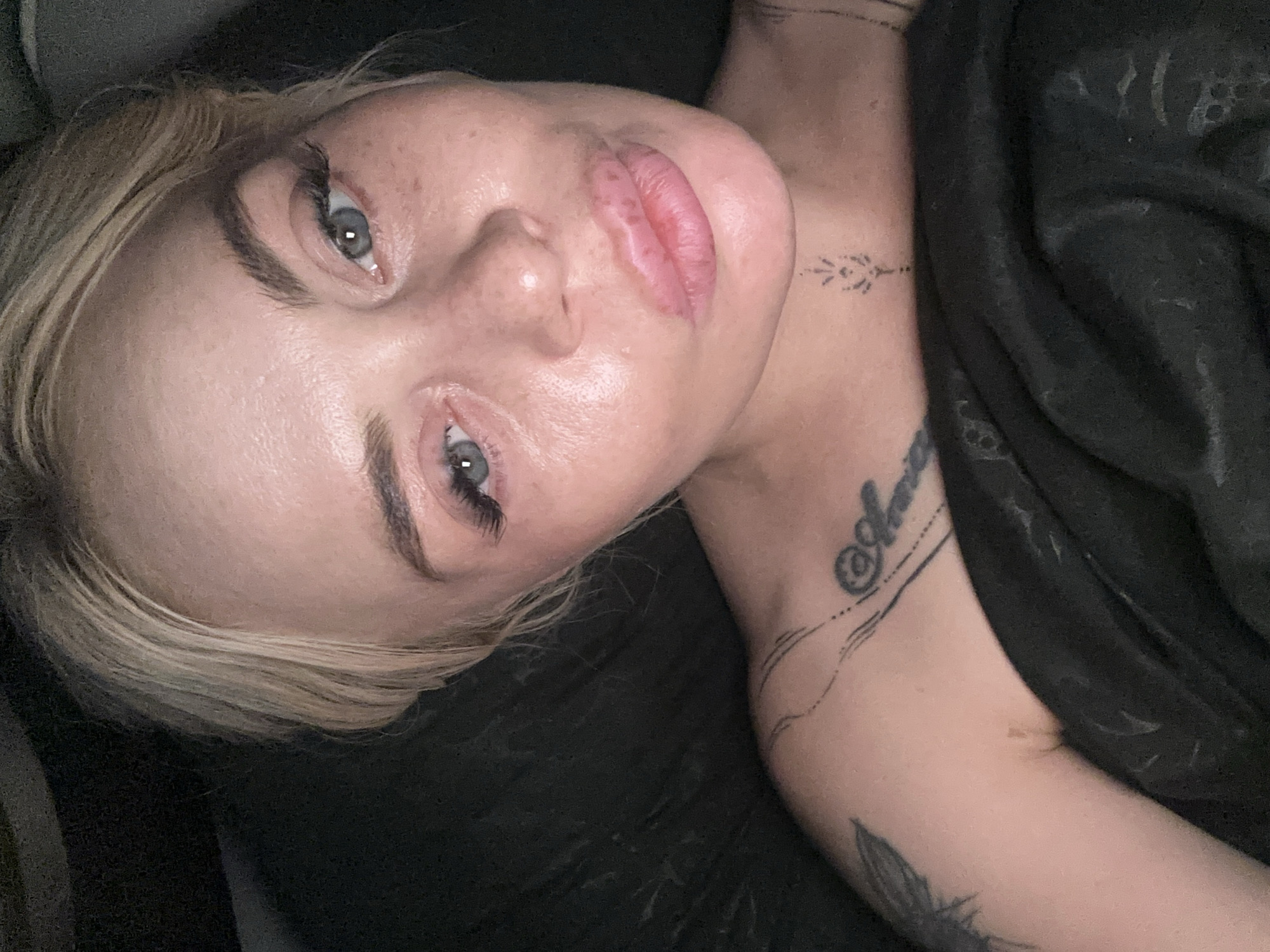 A close-up, overhead shot shows a fair-skinned woman with blonde hair and blue eyes lying down, wrapped in a dark blanket. Her head is tilted slightly upwards, and her lips are pursed. She has on mascara and what appear to be false eyelashes. She has some light freckling on her cheeks and nose. On her upper chest, there are a few tattoos, including one on her left shoulder that appears to be a feather and another on her right chest of what looks like a bird with a cursive script underneath. There is also a small, linear tattoo on her throat. The background is dark and out of focus. The lighting is soft, casting some highlights on her skin.