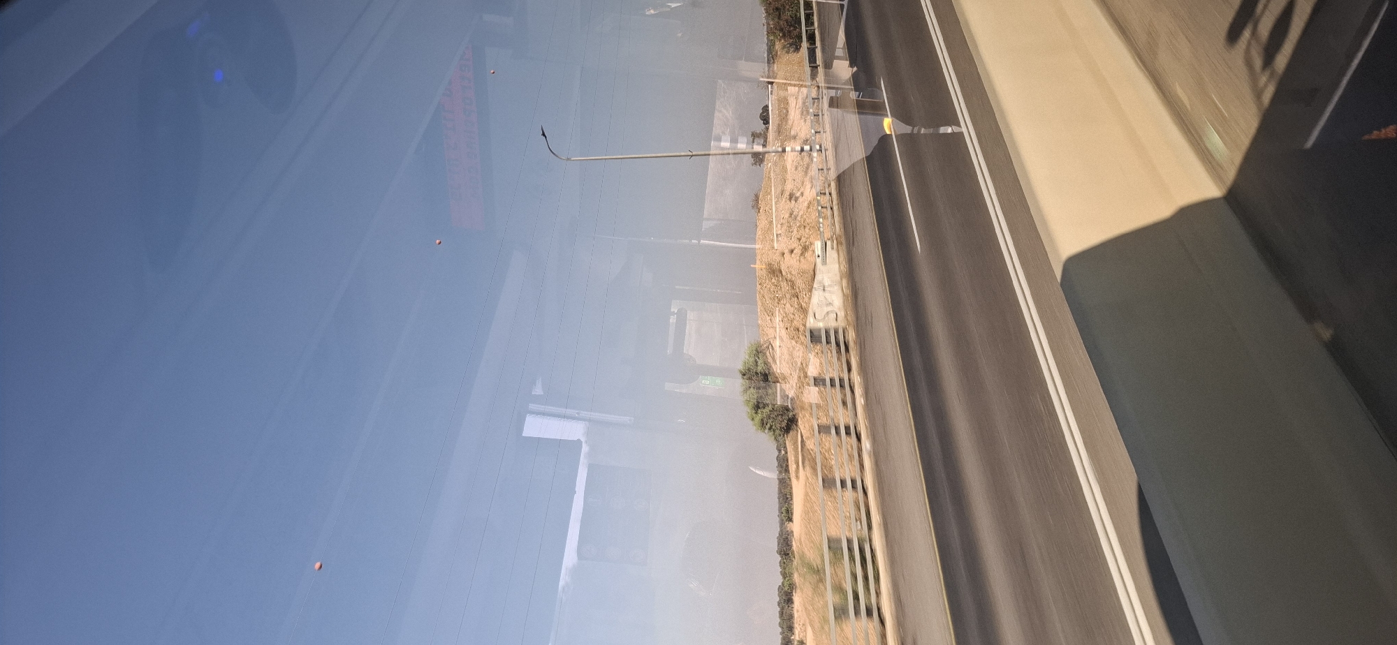 The image captures a daytime view through a reflective surface, likely a window, of a road in the Bnei Shimon Regional Council, Israel. The foreground features a beige concrete barrier with a shadow cast upon it, indicating sunlight from the right. Beyond the barrier is a multi-lane asphalt road with white lane markings, showing motion blur. A metal railing with vertical posts separates the road from a sandy embankment. On the embankment, there are sparse trees and bushes, with evidence of construction or earthworks. A street light stands to the right of the embankment. The sky is clear blue with several small, orange objects, possibly balloons or kites, visible. Reflections on the window obscure parts of the scene, showing abstract shapes and what appears to be text on an illuminated sign. The overall impression is a roadside environment in a semi-arid region under a bright sky.