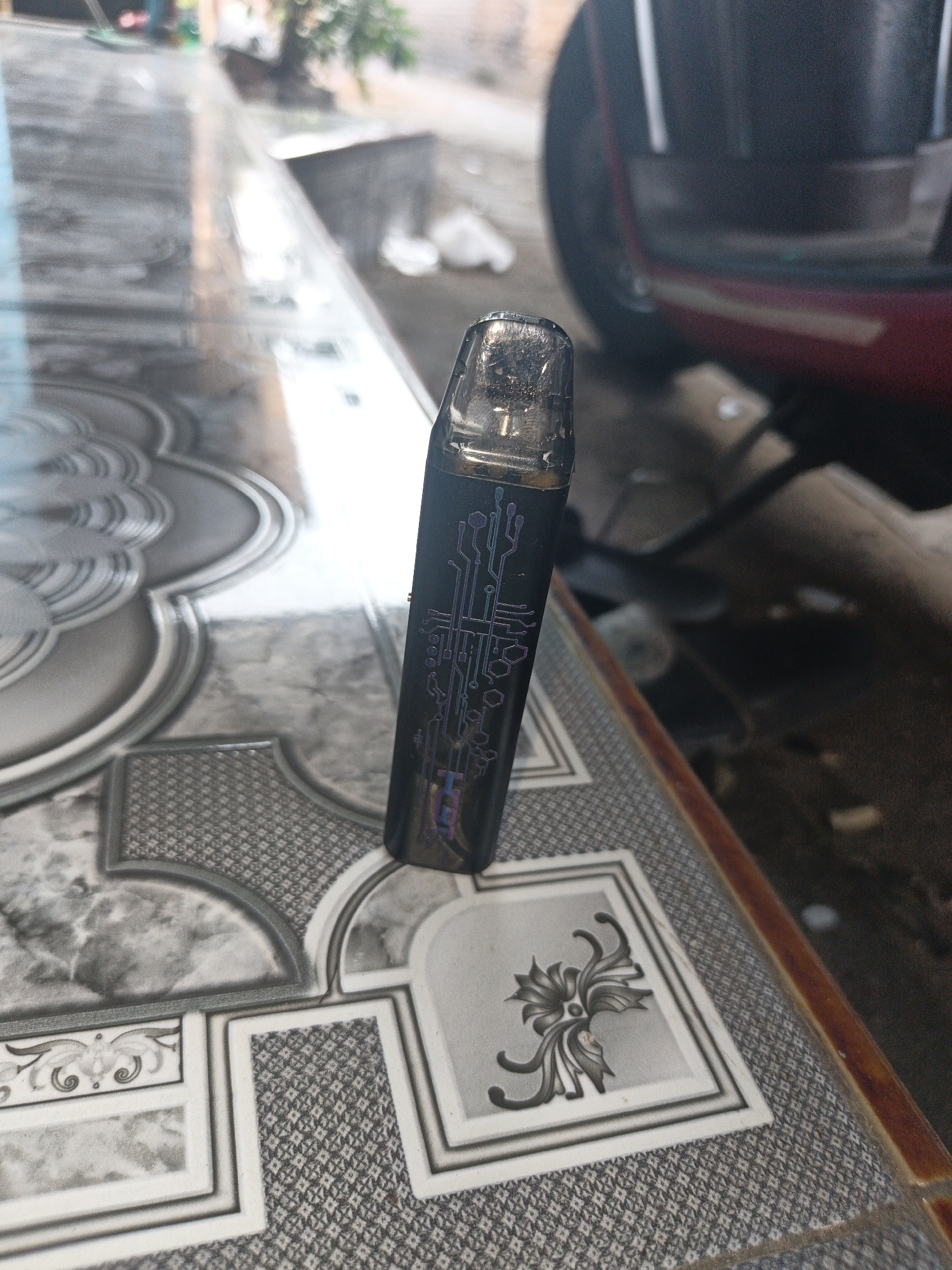 A black vape pen with a circuit board design is on a patterned tabletop. The tabletop has a glossy finish and is divided into sections with grey and white intricate designs. In the background, a red scooter is partially visible. The environment appears to be outdoors, with natural light suggesting a daytime scene. The location is identified as Temuwulan, Indonesia.