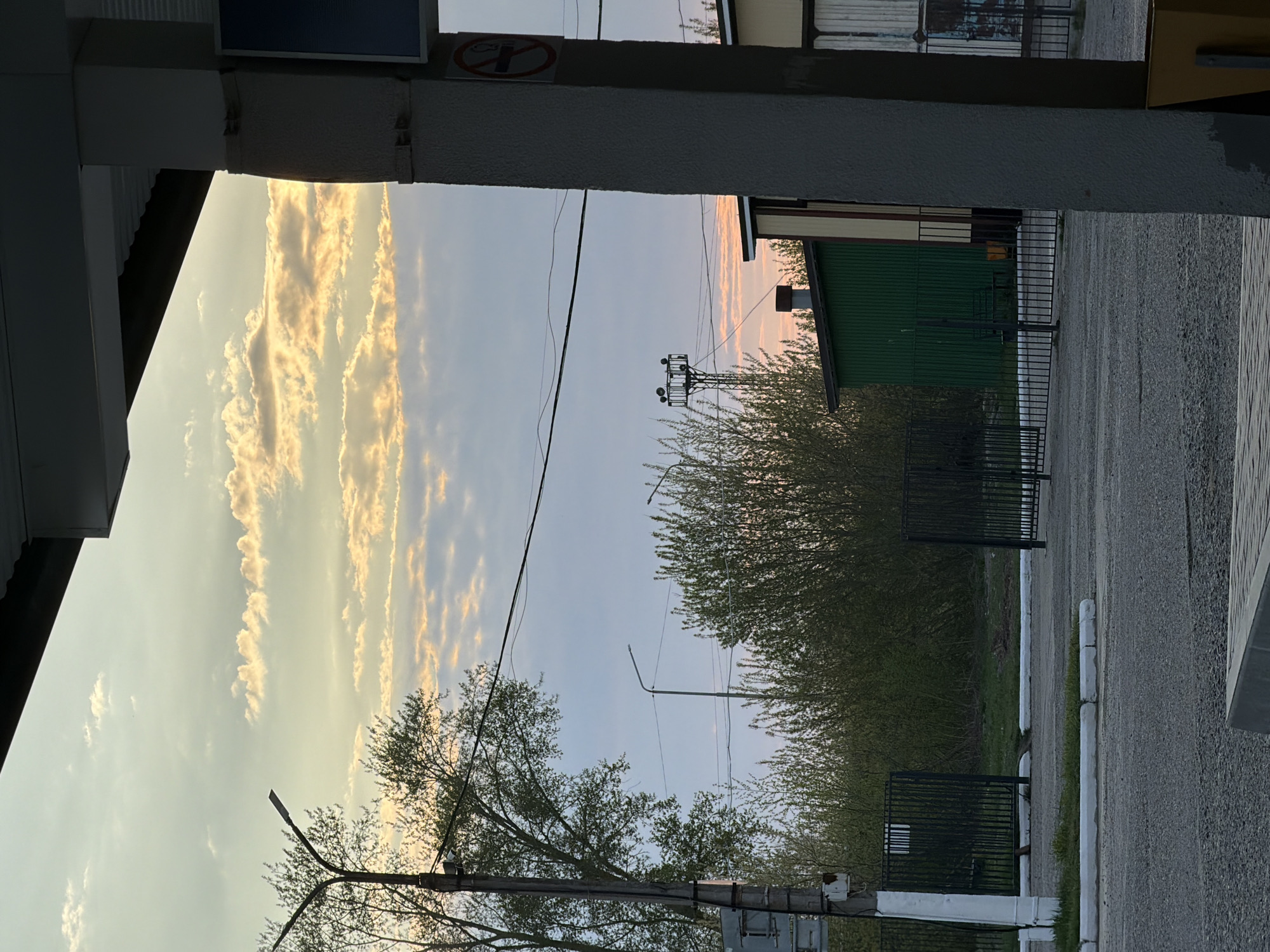 The image shows a street scene in Chaikovsky, Russia. The view is partially framed by architectural elements, possibly a building or overpass structure. The sky is visible, displaying elongated clouds illuminated by the setting sun. A utility pole and power lines run across the sky. Below, trees and dense bushes line the road. A green corrugated iron building is also visible behind a fence. The ground consists of an asphalt or paved surface. No people are visible in the image.