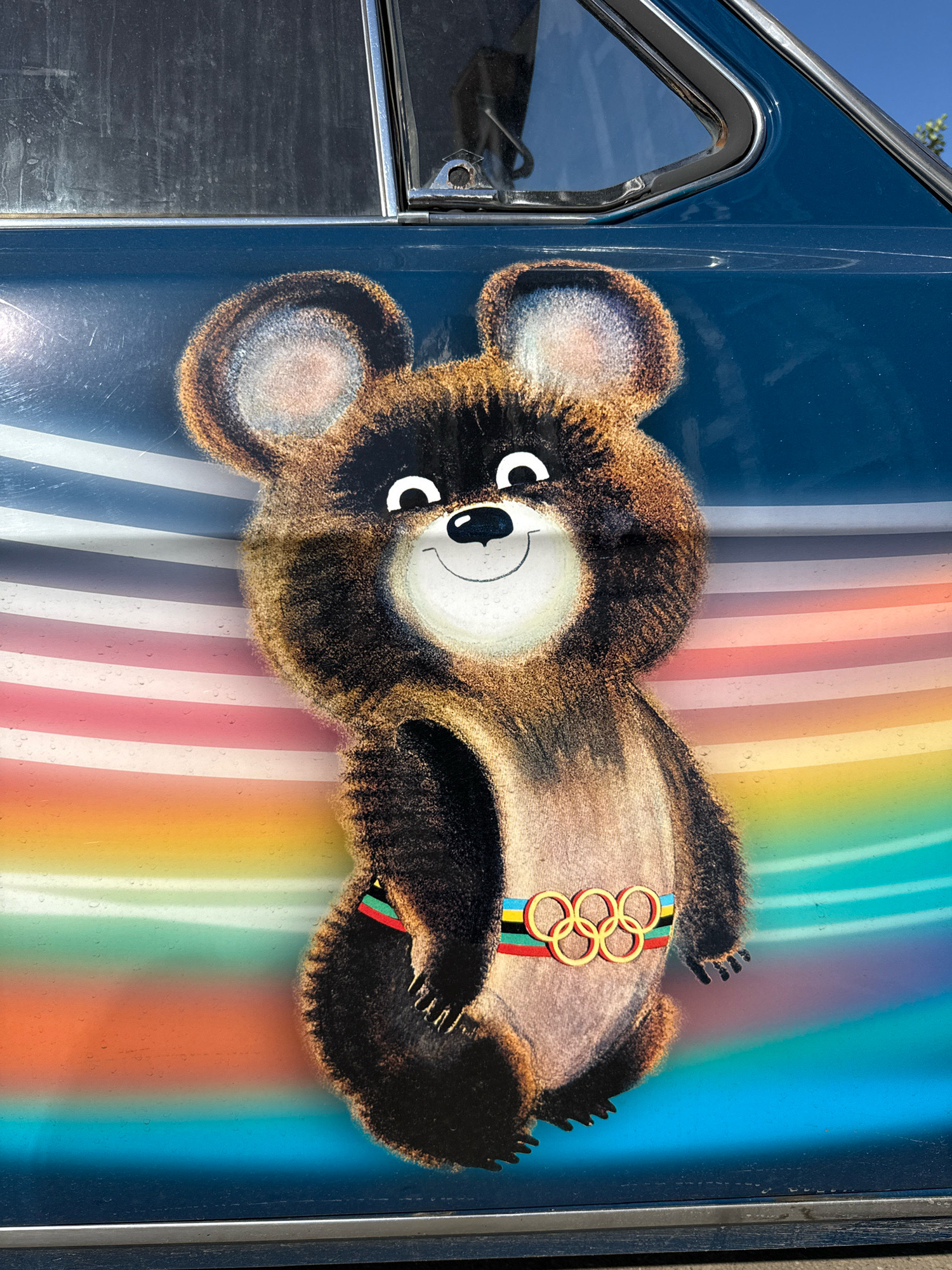 The image is a close-up, exterior shot of a dark blue vehicle, possibly a car, adorned with a painted mural of Misha, the mascot of the 1980 Moscow Olympics. Misha is depicted as a cartoon bear with brown fur, large ears, and a friendly smile, wearing a sash with the Olympic rings. The mural is set against a backdrop of rainbow-colored horizontal stripes. The vehicle appears to be parked outdoors, under a clear sky, as evidenced by the sunlight reflecting off its surface and a glimpse of blue sky at the top right of the image. The vehicle's window and chrome trim are visible. The location is specified as the city of Rusanovo, Russia.