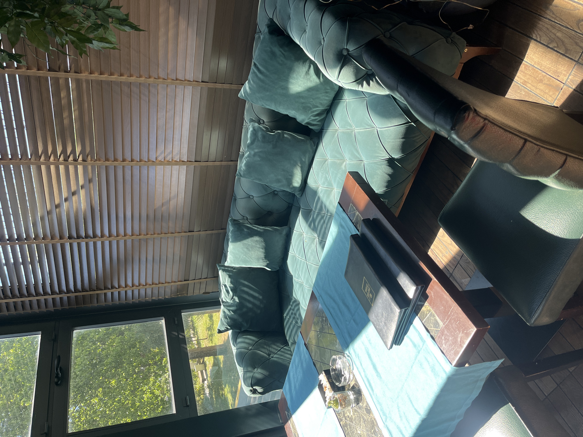 The image shows an indoor seating area, possibly in a cafe or restaurant. A plush, teal-colored tufted sofa with throw pillows sits against a window-lined wall offering a view of trees and greenery outside. A small table in front of the sofa is covered with a light blue runner and holds menus and glassware. A chair is positioned near the table, suggesting a dining or conversational setting. The ceiling features slatted blinds allowing natural light to filter into the space. Based on the architectural style, furnishings, and possible view, the location could potentially be in the city of Budva, Montenegro.