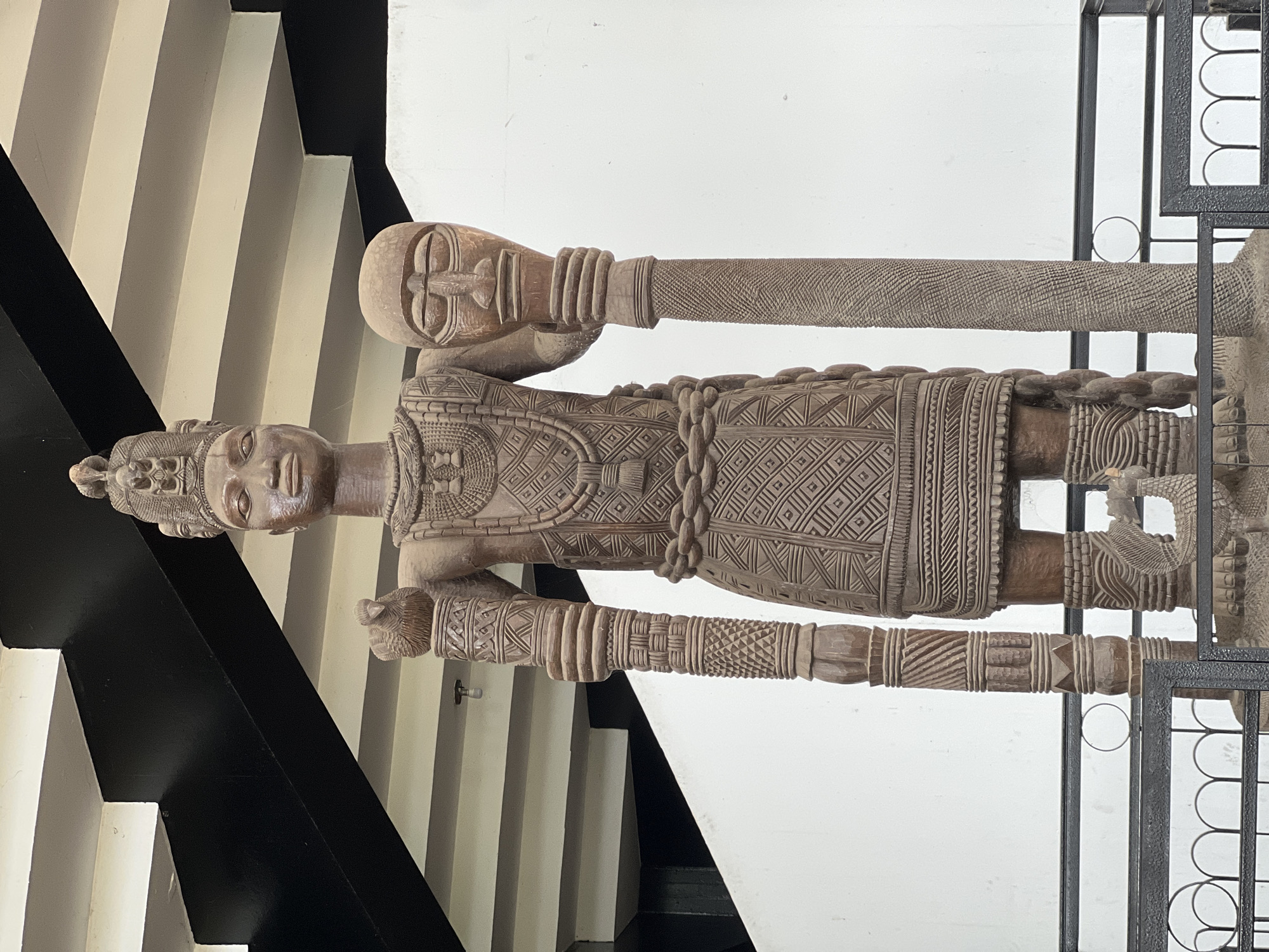This is a full-body, eye-level photograph of a large, intricately carved wooden sculpture of a regal figure, likely an Oba (king) of the Yoruba people. The sculpture stands in what appears to be an indoor exhibition space. The figure is depicted standing upright, facing the viewer, and is adorned with a tall, elaborate headdress featuring multiple smaller heads. It wears a detailed necklace and a patterned tunic or robe. In its right hand, the figure holds a bird sculpture, and in its left hand, it holds a staff topped with a human head. The sculpture exhibits rich textural detail, with patterns carved into its clothing, arms, and the staff.
Behind the sculpture, a white wall serves as the backdrop, with a black and white patterned ceiling structure above. The sculpture is positioned in front of a metal barrier, suggesting it is an exhibit. The overall lighting is bright, illuminating the sculpture and its surroundings. While the image captures a sculpture, its depiction and the context suggest a connection to the historical and artistic heritage of Ife, Nigeria, a city renowned for its ancient Ife bronze and terracotta sculptures, as well as its wooden carvings.