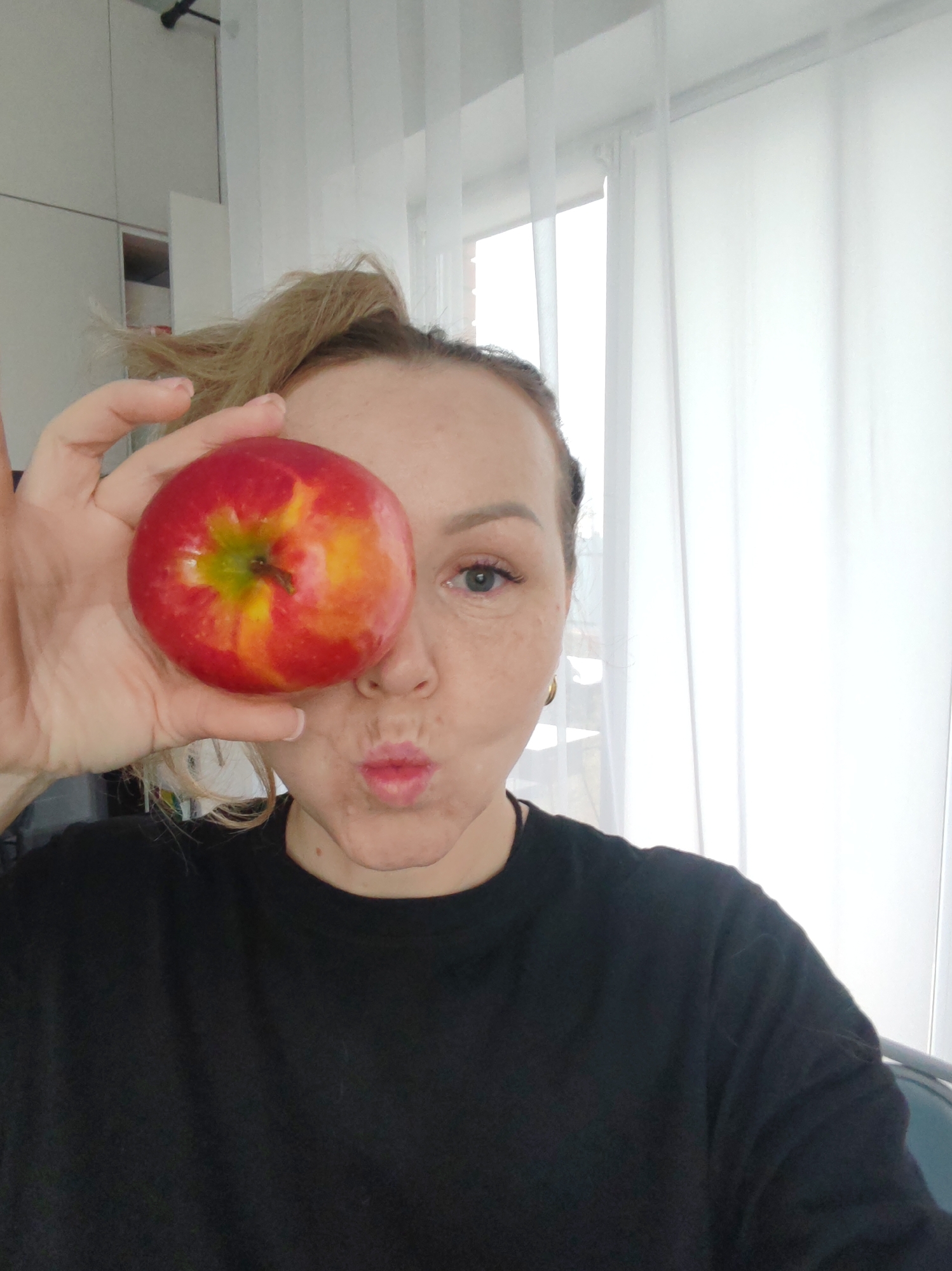 A woman with blond hair in a ponytail is holding a red apple in front of her face, covering one eye. She is wearing a black t-shirt. The background shows a room with white curtains and what appears to be kitchen cabinets. This image does not contain sufficient information to confirm the specific location in Chaikovsky, Russia.