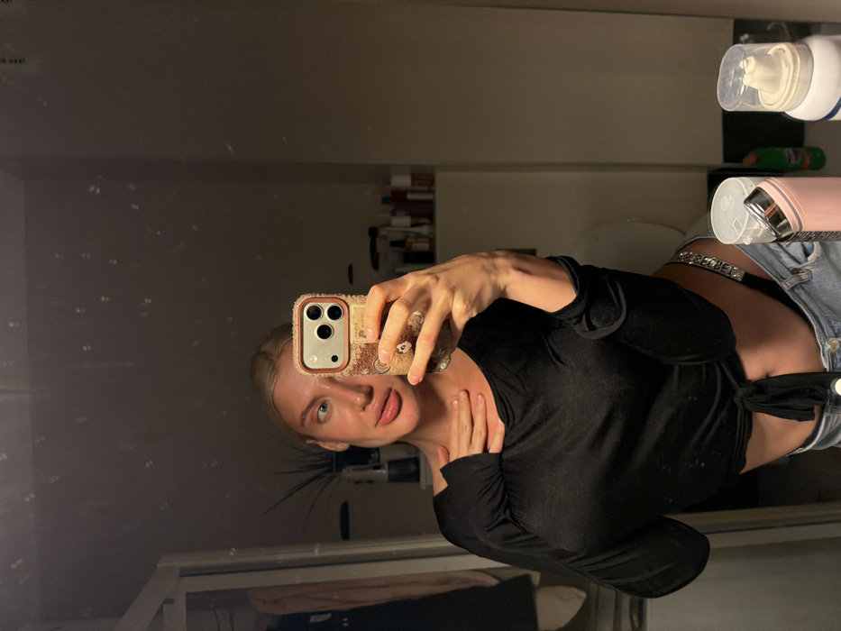 The image displays a mirror selfie of a blonde-haired individual, likely female, captured indoors in a dimly lit room, consistent with a private residential or hotel bathroom. The subject wears a black long-sleeved crop top, tied at the waist, exposing a portion of the abdomen, and light-colored denim jeans. A rhinestone-embellished strap is visible on the left hip above the jeans' waistband. A silver ring is present on the left ring finger. The subject holds a silver iPhone, equipped with a multi-camera system and housed in a light brown textured case, in the right hand. The left hand rests on the upper chest/collarbone area. The facial expression includes pursed lips and eyes directed to the right. The reflected background reveals a plant in a pot and a dark hanging garment to the left, and shelves containing various toiletries and a partially visible white toilet lid to the right. In the foreground, on a surface in front of the mirror, a pink insulated beverage bottle and a white aerosol can are visible. No features within the image provide specific information to identify a location in Budva, Montenegro.