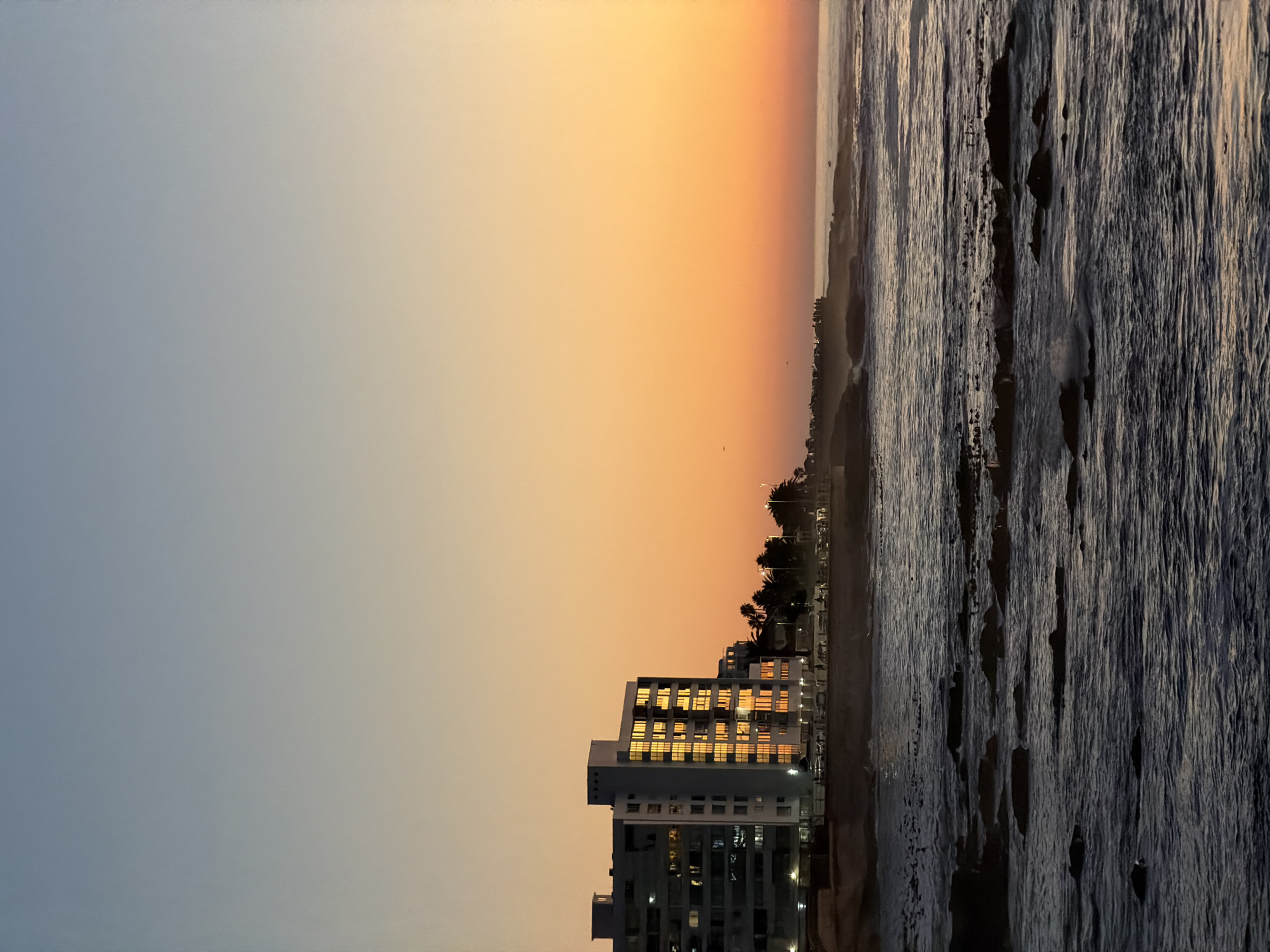 A serene view captures a tall building along the coastline under a sky transitioning from dusky blue to warm sunset hues, adjacent to the textured waters of the ocean. The building's windows reflect the golden light, suggesting activity within. Palm trees and street lights line the beach area, indicating a developed coastal location. The photograph appears to have been taken in a coastal city, similar to Cape Town, South Africa. The waves crashing against the shoreline are faintly visible, adding depth to the scene.