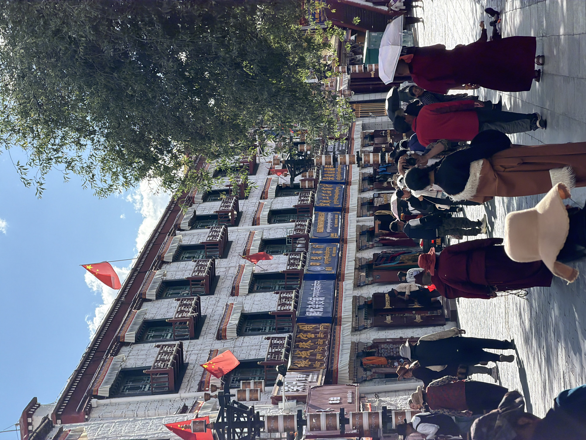 This image depicts a bustling street scene in a cultural or historic district, likely in Lhasa, China, given the location context of Chengguan District. The focal point is a multi-story building characterized by its white, textured walls, dark-framed windows, and ornate wooden balconies or decorative elements. Several Chinese national flags, red with five yellow stars, are prominently displayed along the building's facade.
The street level is filled with numerous people, indicating a busy atmosphere. Individuals are seen walking, standing, and possibly shopping. Some people are dressed in long, traditional robes, while others wear casual modern attire. Several people are holding umbrellas, presumably for sun protection under the bright, clear sky. The strong sunlight casts visible shadows on the paved ground, suggesting it is midday or early afternoon.
Along the ground floor of the building, numerous shops or stalls are evident, featuring a variety of goods and decorative elements. There are multiple signs, some blue with white or yellow Chinese characters, and others brown with gold lettering. Visible text on these signs includes "拉萨市民族传统工艺品..." (Lhasa City National Traditional Handicrafts...) and mentions of "唐卡" (Thangka) and "画廊" (Gallery), suggesting businesses focused on traditional arts and crafts. Below the shops, what appear to be prayer wheels or similar religious artifacts are visible. A large, leafy green tree partially shades the top left of the scene, its foliage contrasting with the clear blue sky and scattered white clouds. The overall scene conveys a vibrant blend of daily life, commerce, and traditional culture.