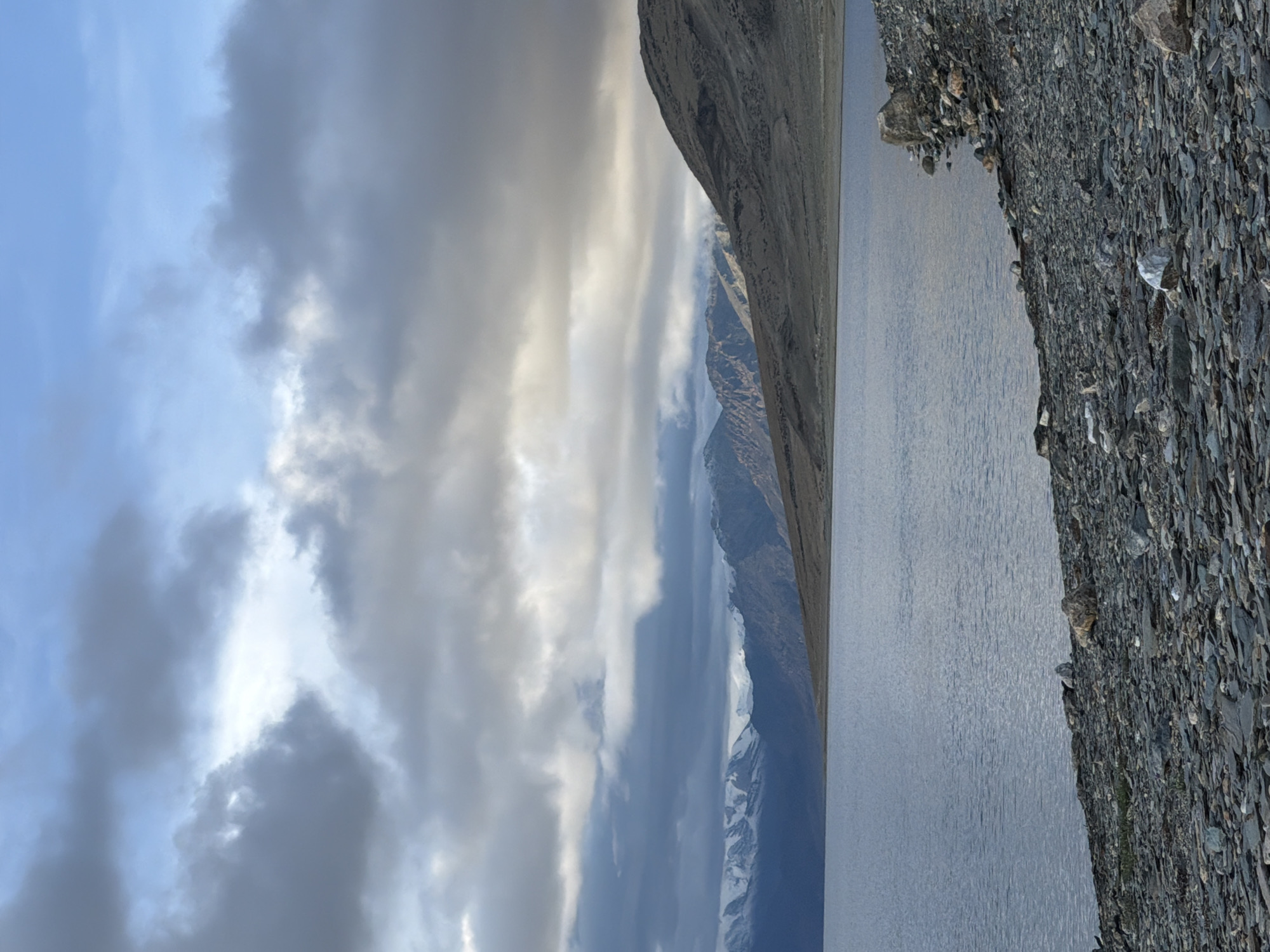 A vertical, medium shot captures a vast, serene lake with a rocky shoreline under a dramatic, cloudy sky. In the distance, rugged, snow-capped mountains rise against the horizon, their peaks catching the soft light breaking through the clouds. The lake's surface is calm, reflecting the muted tones of the sky and mountains. The shoreline is covered in dark, grey stones and pebbles, with a few larger rocks scattered amongst them. The sky is a blend of blues and greys, with some areas illuminated by sunlight, creating a sense of depth and atmosphere. The overall scene conveys a feeling of vastness and natural beauty, characteristic of a high-altitude landscape in Ladakh, India.