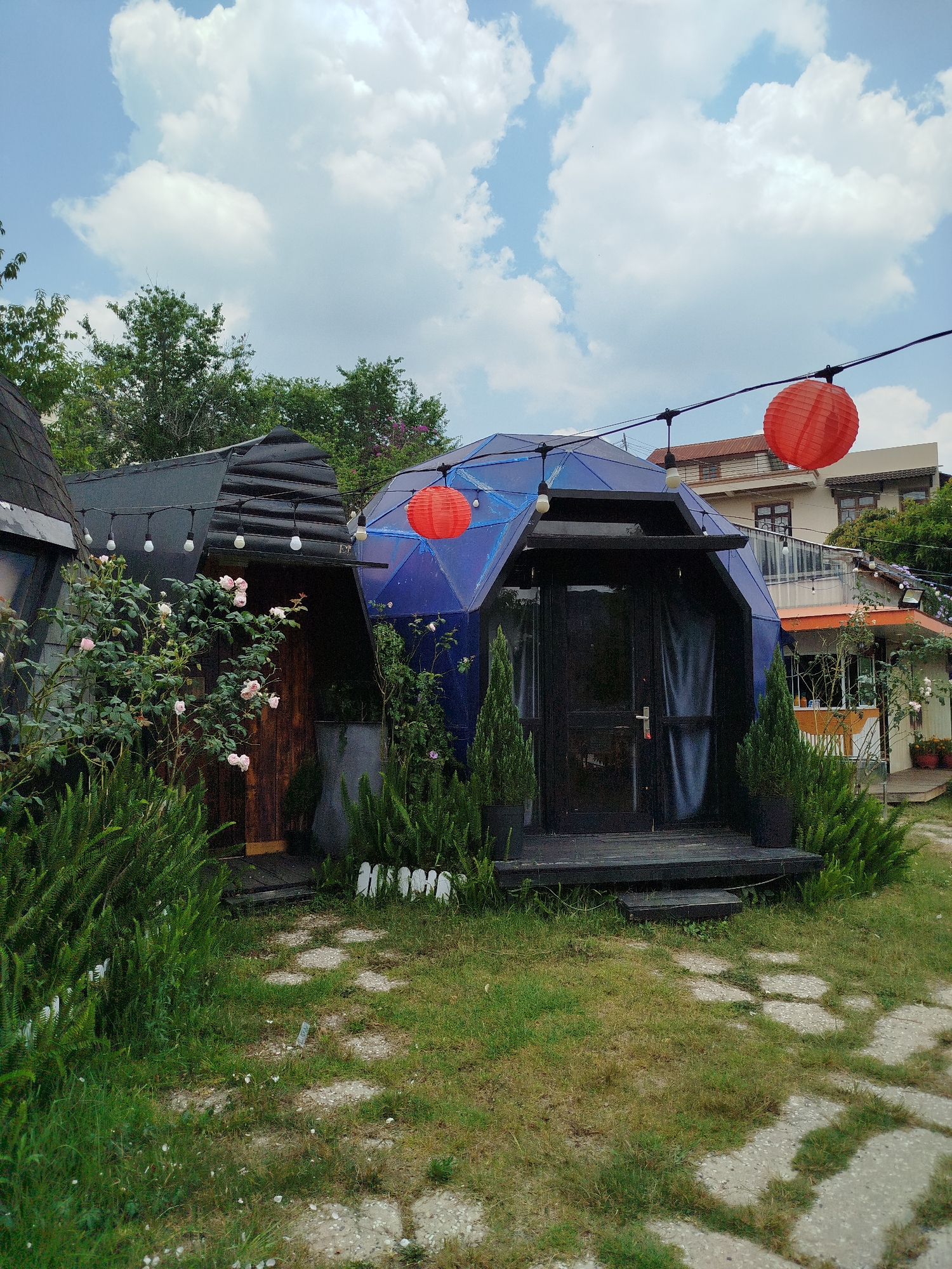 A daytime exterior shot shows a small, dome-shaped, cobalt-blue structure with a black door and frame. A matching black porch with two steps is at the entrance, flanked by small, dark green conifers in black planters. A pathway composed of light grey irregular stone shapes is visible in the foreground. To the left is another smaller, dark-roofed structure with wooden siding, partially obscured by dense greenery, including a rose bush with light pink roses. A string of round lights hangs from the eaves of both structures. Above and to the right, red paper lanterns are strung between the structures and a two-story building with an orange awning and a balcony. Trees and a cloudy blue sky are visible in the background. The photo's location is Da Lat, Vietnam, implied by the architectural style and landscaping.