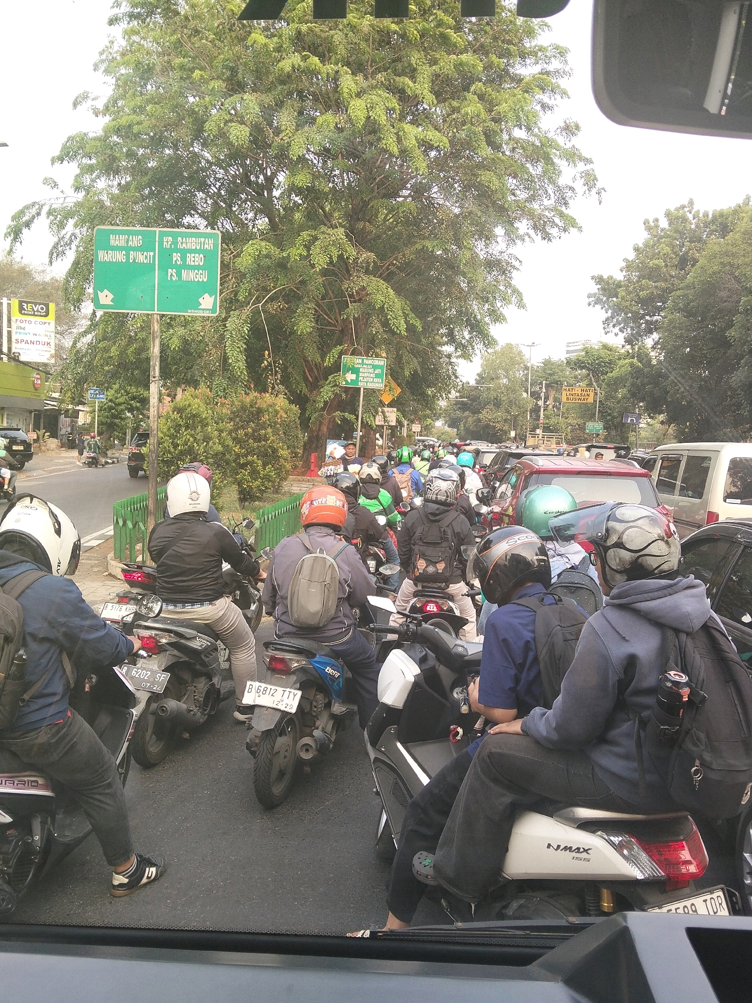 In South Jakarta, Indonesia, a traffic jam is depicted on a road lined with trees and buildings. The foreground shows several people riding motorcycles, some wearing helmets and backpacks. One person in the foreground is wearing a black jacket, gray pants, and black and white sneakers. The middle ground features more motorcycles and cars, including a red SUV and a silver van. A green signpost with Indonesian text is visible, indicating directions to "MAMPANG, WARUNG BUNCIT," "KP. RAMBUTAN, PS. REBO, PS. MINGGU." Another yellow sign displays a warning about busway traffic. The overall scene suggests a busy urban environment with significant vehicular congestion.