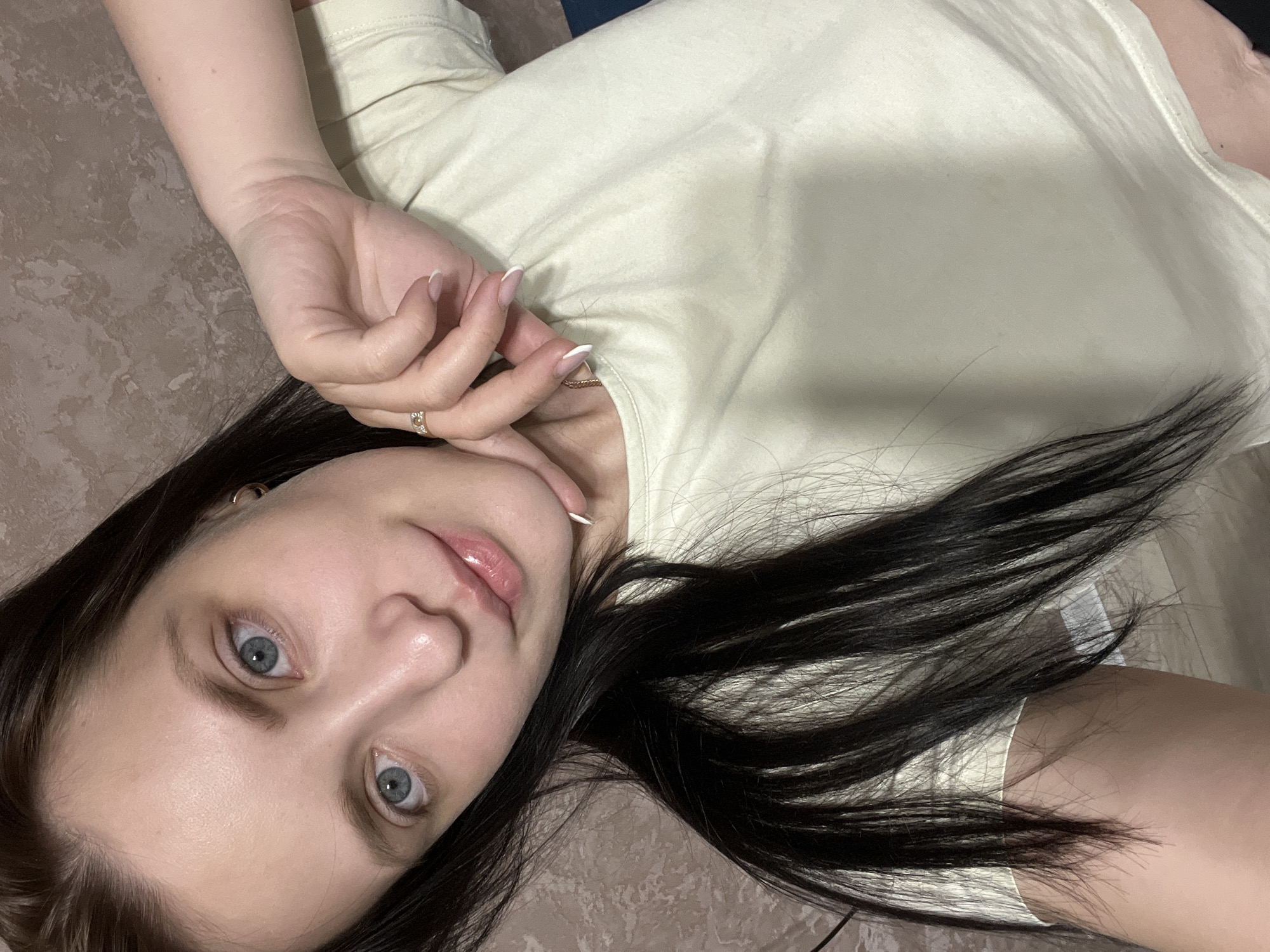 The image depicts a woman with dark hair and light eyes lying down. Her head is tilted, and she is looking directly at the camera. She is wearing a light-colored T-shirt. Her right hand is near her face. The background is a mottled brown surface. The location cannot be determined from the image and is unlikely to be identified as Марковский, Russia.