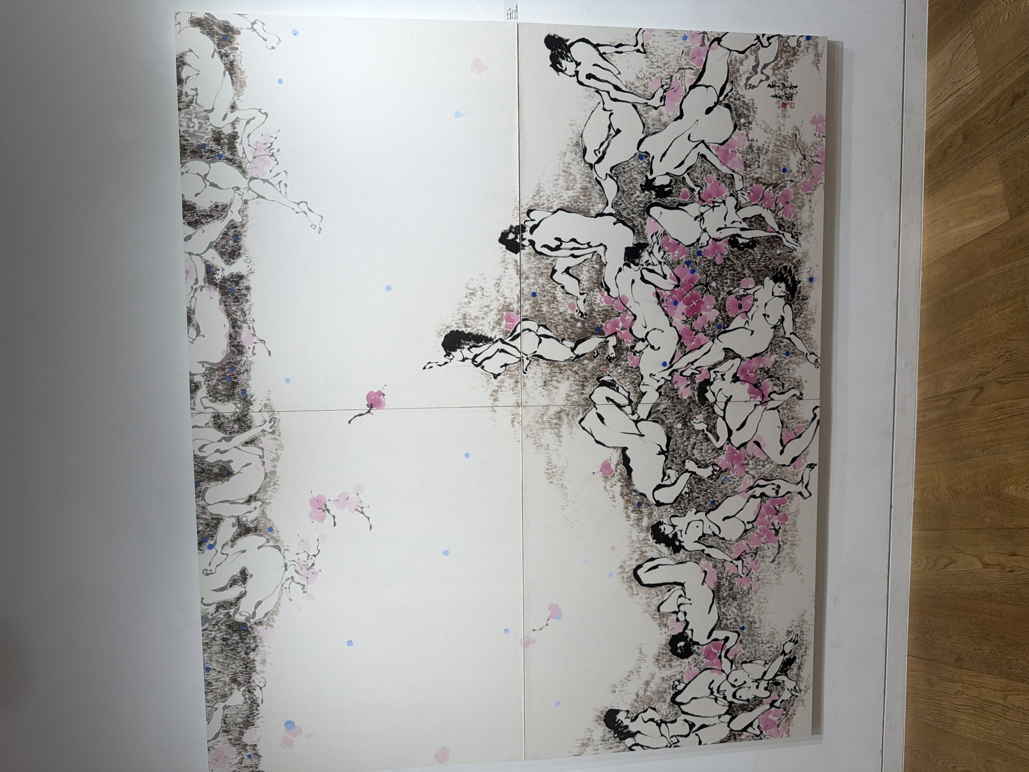 The image shows a large, multi-panel ink wash painting of numerous unclothed figures in various reclining and standing poses. The figures are rendered with simple, fluid lines and are primarily depicted in black ink with washes of gray to suggest shading. Scattered throughout the composition are delicate pink blossoms and small blue dots, which appear to be part of the artistic style rather than depicting specific objects. The background is largely white, with some areas of textured brown ink suggesting ground or foliage. In the bottom right corner of the artwork, there is a red artist's seal and what appears to be Chinese or Korean calligraphy. The painting is displayed on a white wall above a wooden floor. A white label with text is affixed to the wall to the right of the painting. The image appears to have been taken in a gallery or exhibition space. Based on the information provided, the location is the K-POP Gallery in Seoul, South Korea.