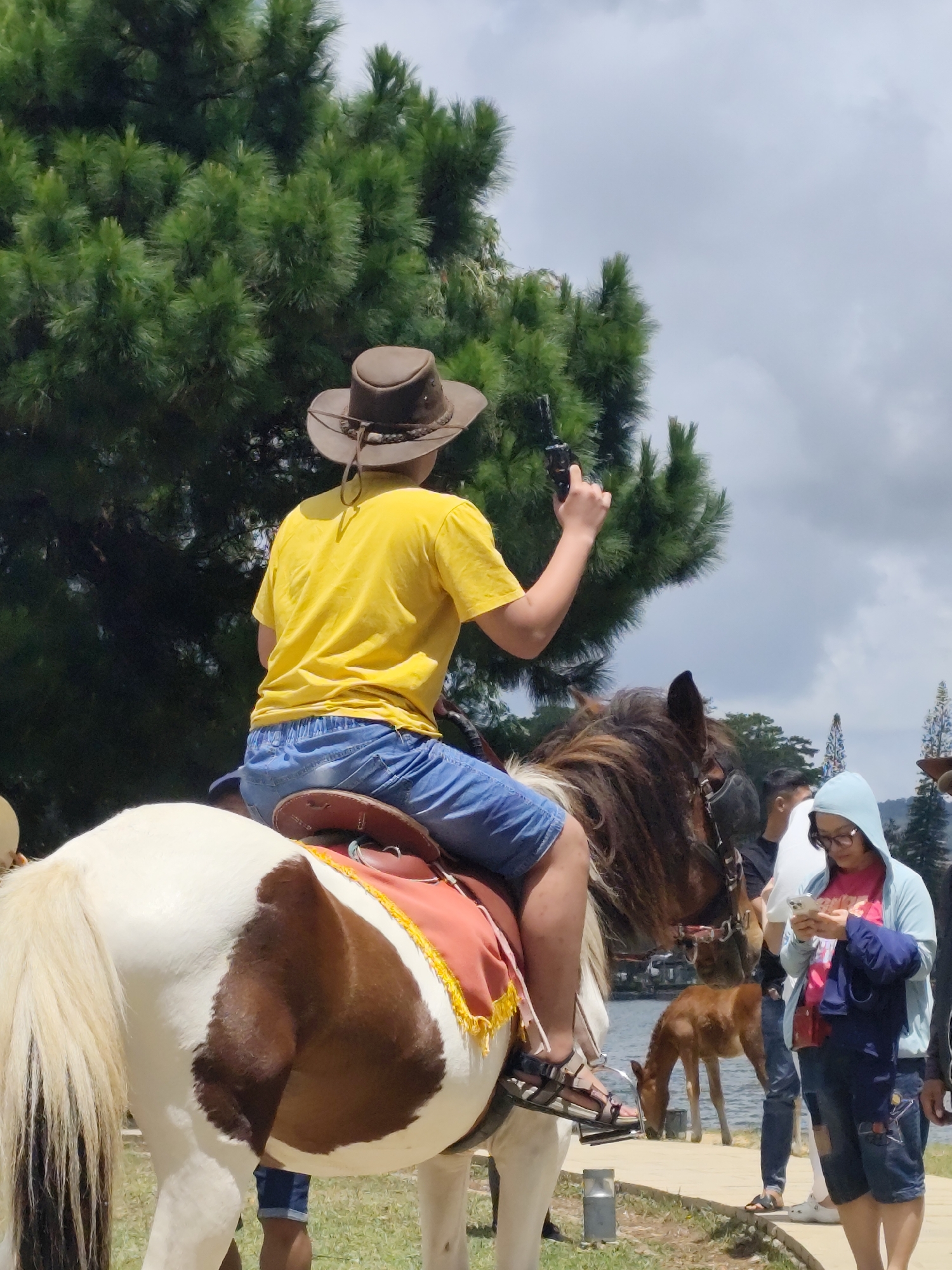 A person wearing a cowboy hat, yellow t-shirt, and blue shorts is riding a pinto horse. The rider is holding a dark-colored object, possibly a toy gun, in their right hand. The horse has a brown saddle blanket with yellow fringe. In the background, there are pine trees, a body of water, and other people. One person, wearing a blue hoodie and glasses, is looking at their phone. A small brown foal is drinking water at the edge of the body of water. The scene appears to be a tourist attraction in Da Lat, Vietnam, likely a horse riding facility.