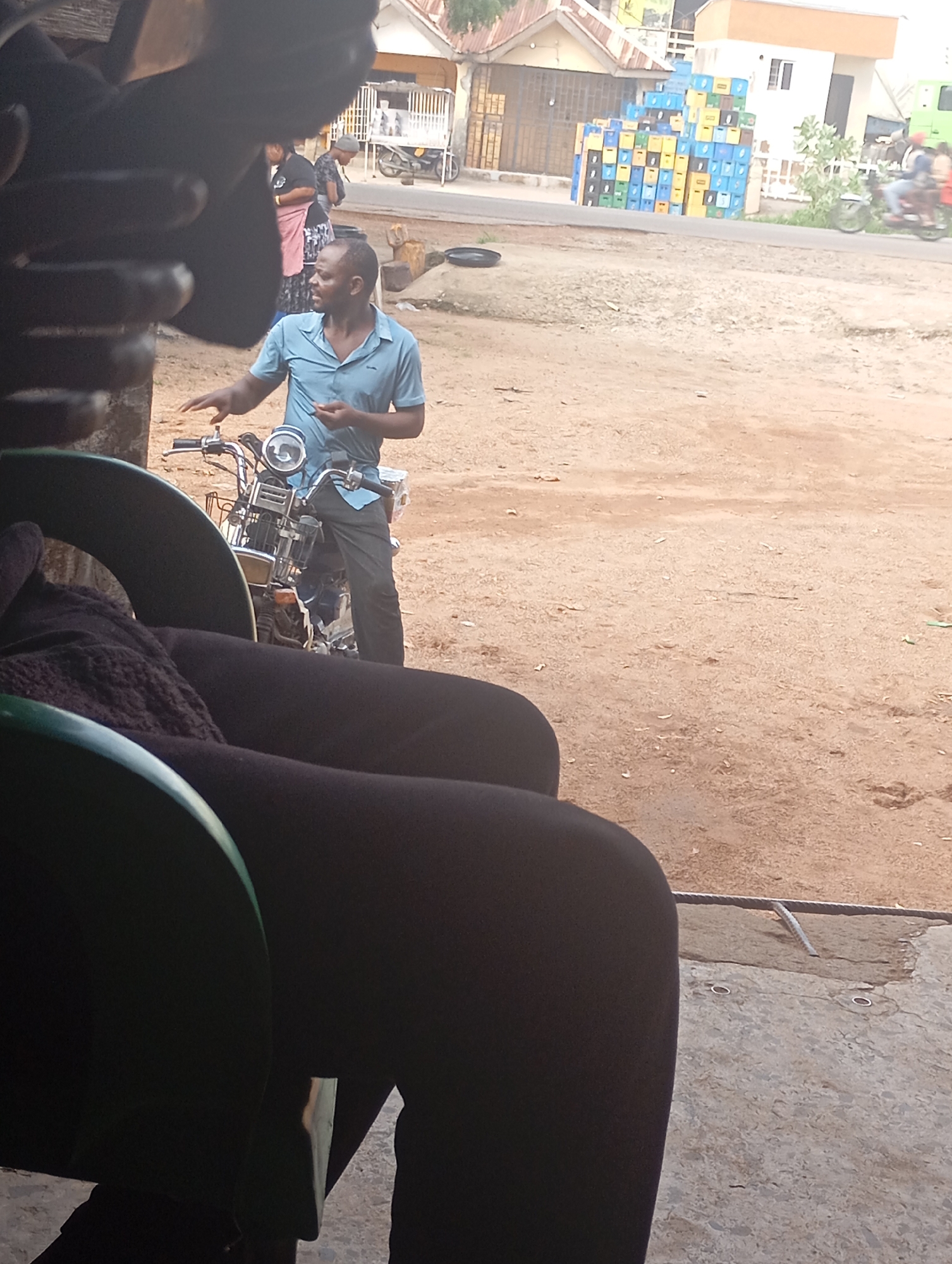 In Shendam, Nigeria, a man in a blue polo shirt and dark pants stands beside a motorcycle. He appears to be in motion, possibly gesturing with his right hand. The motorcycle is parked on a dusty, unpaved surface. In the background, a stack of colorful plastic crates is visible, along with buildings and what appears to be a road with a motorcycle and two people riding on it. The overall scene suggests an outdoor marketplace or a street in a Nigerian town.