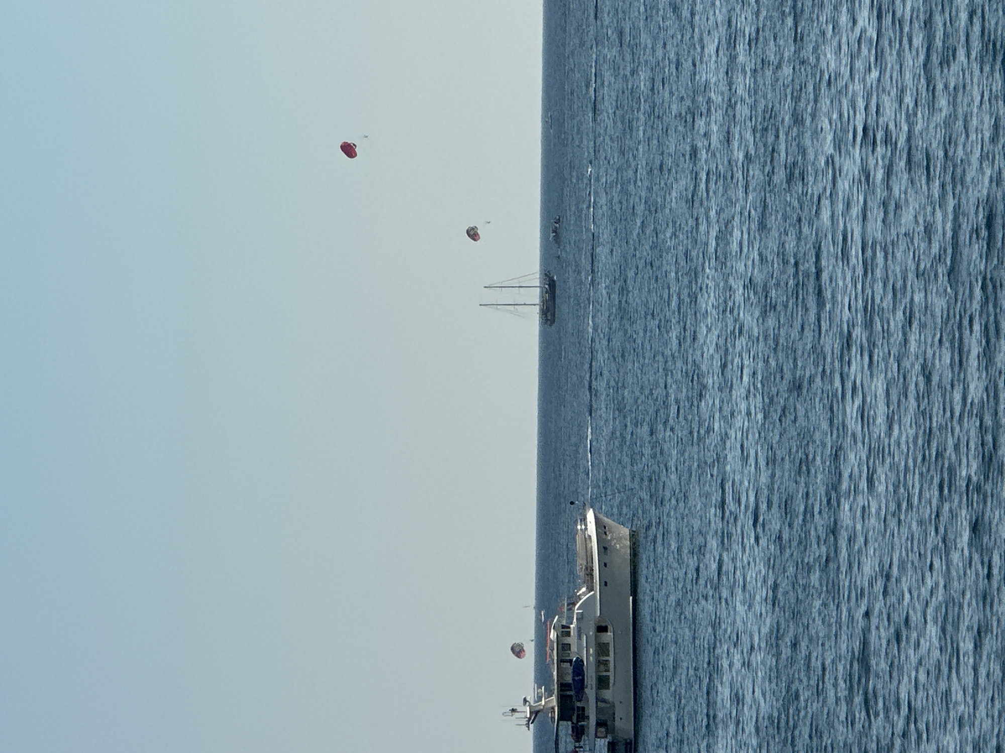 An expansive, calm blue sea under a pale blue sky forms the backdrop for a leisure scene. In the foreground, a white yacht with multiple decks is positioned to the left, its hull reflecting the light. Further in the distance, a two-masted sailboat is visible. Three paragliders are in the air. One is a bright red, another is a mix of red and black, and a third, smaller red paraglider is also present. The overall impression is one of a sunny day at sea, likely near a coastal area known for water sports. This scene is consistent with the popular tourist destinations along the Budva Riviera in Montenegro, where such activities are common.